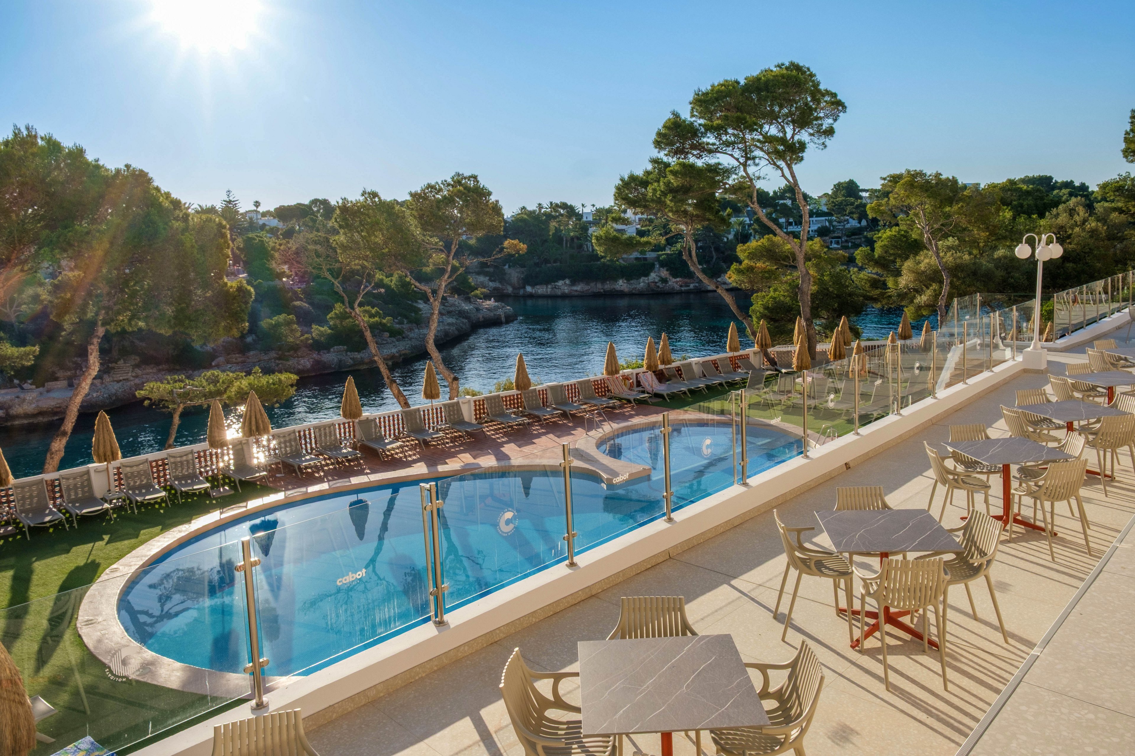 An elevated terrace with tables and chairs overlooks a curved outdoor swimming pool surrounded by sun loungers and umbrellas. Beyond the pool, a calm bay is bordered by rocky shores and tall pine trees under a clear blue sky. Glass railings line the terrace, enhancing the open, airy feel while maintaining safety. The bright sunlight and warm tones create a relaxed vacation atmosphere at this waterfront resort.