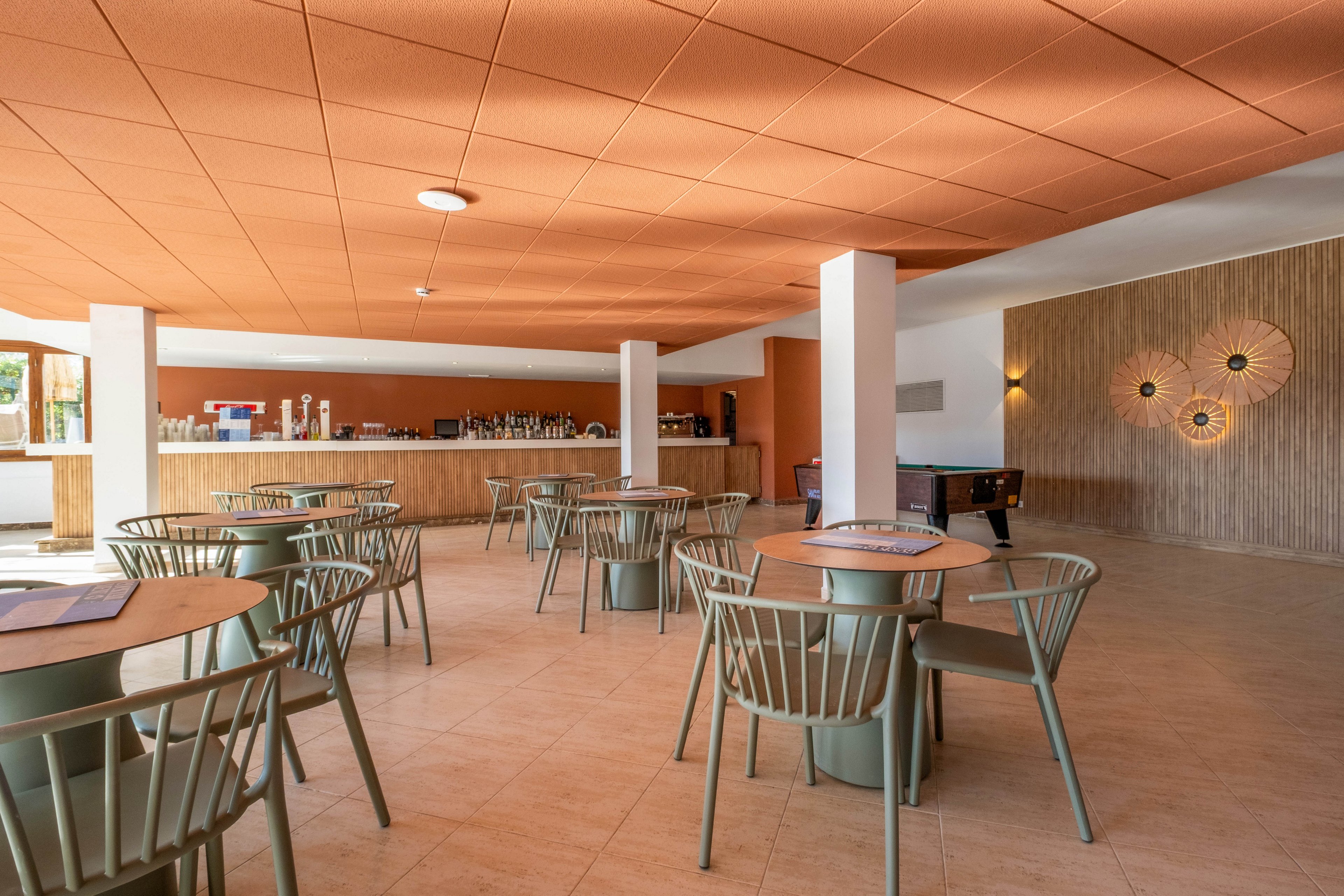 A contemporary bar area features neatly arranged tables and chairs in a spacious room. The setting includes a long counter with drink dispensers and bar equipment along the back wall. Warm earthy tones dominate the space, with an orange ceiling, light tiled floor, and wooden wall panels. Decorative circular wall lights and large windows contribute to a relaxed, inviting atmosphere.
