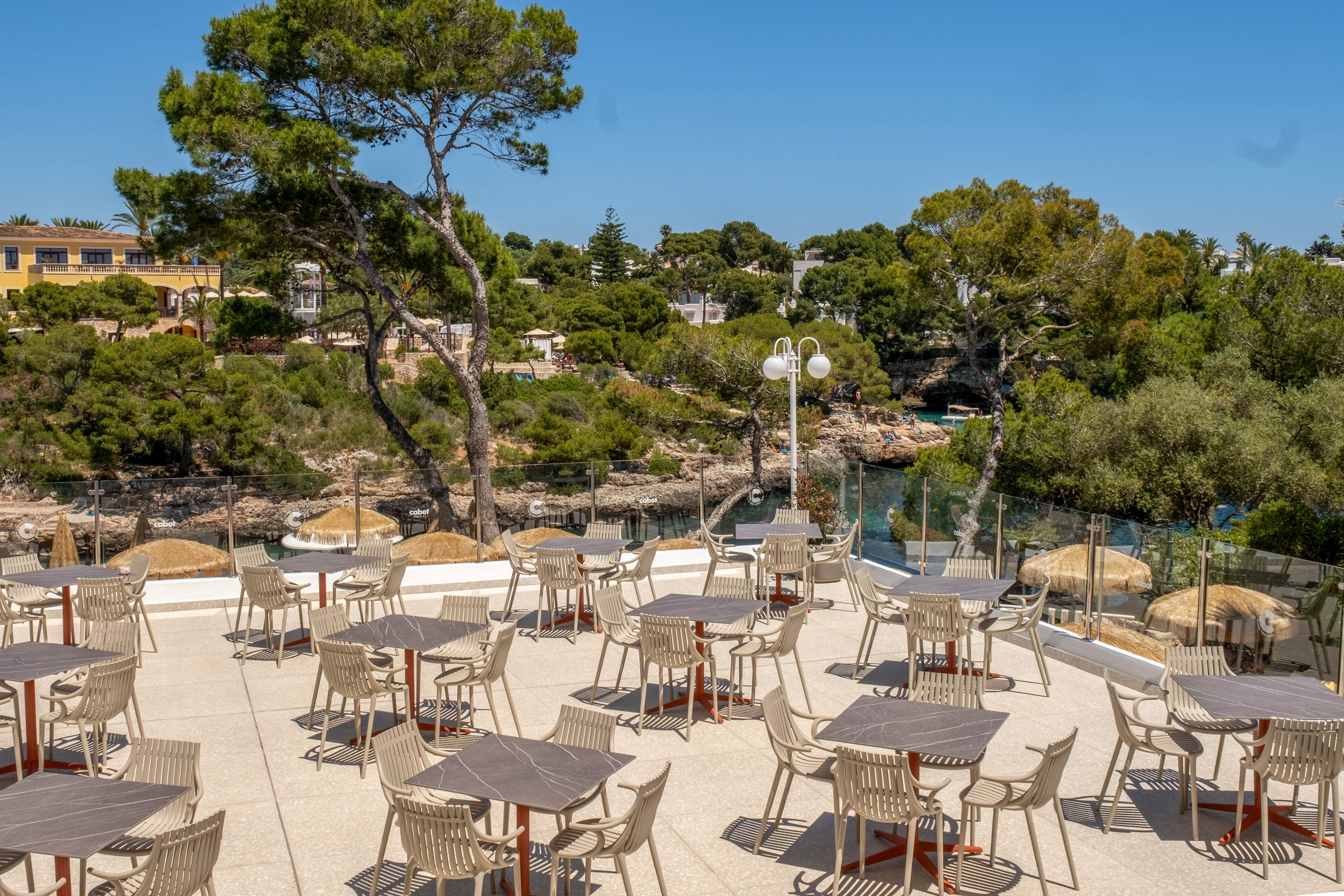 An empty outdoor terrace is set with multiple tables and chairs under bright sunlight. The space overlooks a rocky coastal cove bordered by lush green trees and shrubs. Glass railings line the edge of the terrace, providing an unobstructed view of the water and surrounding landscape. Buildings are visible in the background, suggesting a resort or restaurant setting in a tranquil seaside location.