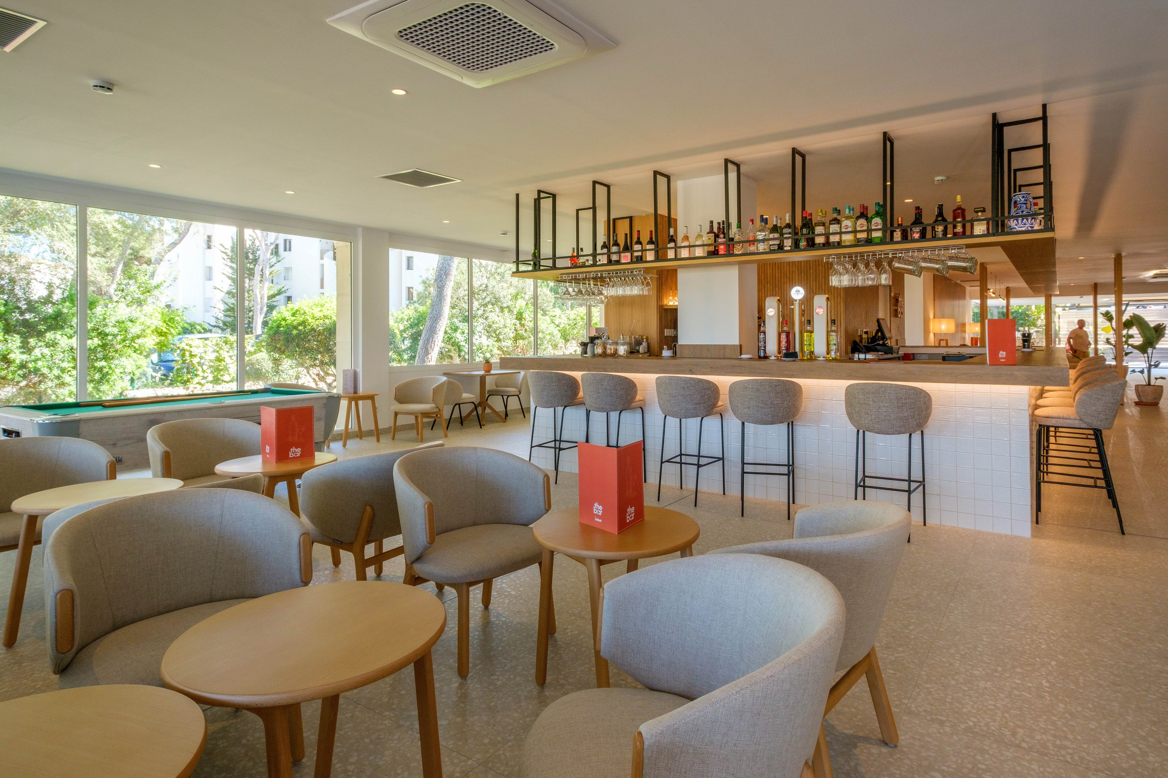 A modern bar lounge features light wood tables and upholstered armchairs arranged in a spacious seating area. The long bar counter with high stools is lined with various bottles and glassware, creating a stylish focal point. Large windows bring in natural light and reveal greenery outside, enhancing the airy atmosphere. Red table tents add a pop of color to the otherwise neutral palette of beige, white, and wood tones.