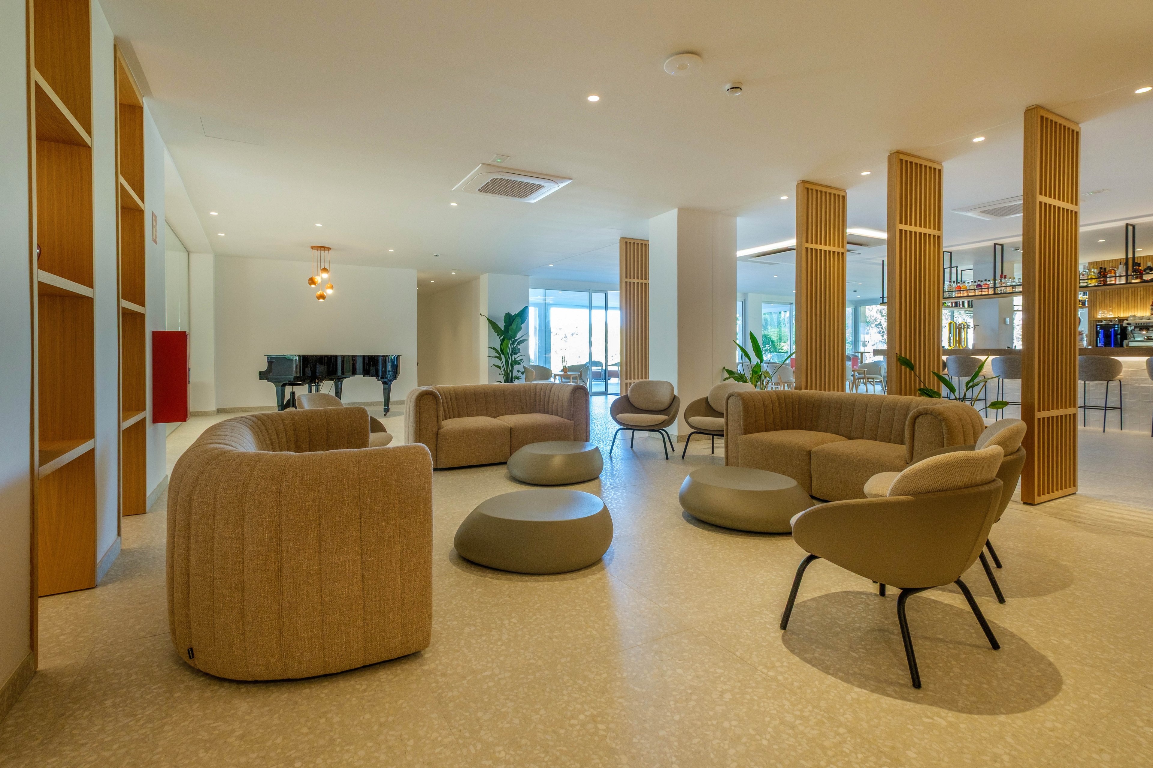 A spacious modern lounge features rounded armchairs and low ottomans arranged for casual conversation. Light wood partitions and neutral-toned flooring create an open, airy atmosphere. A bar area with stools and glass shelving is visible in the background, alongside large windows bringing in natural light. A black grand piano stands near the windows, adding an elegant focal point to the room.