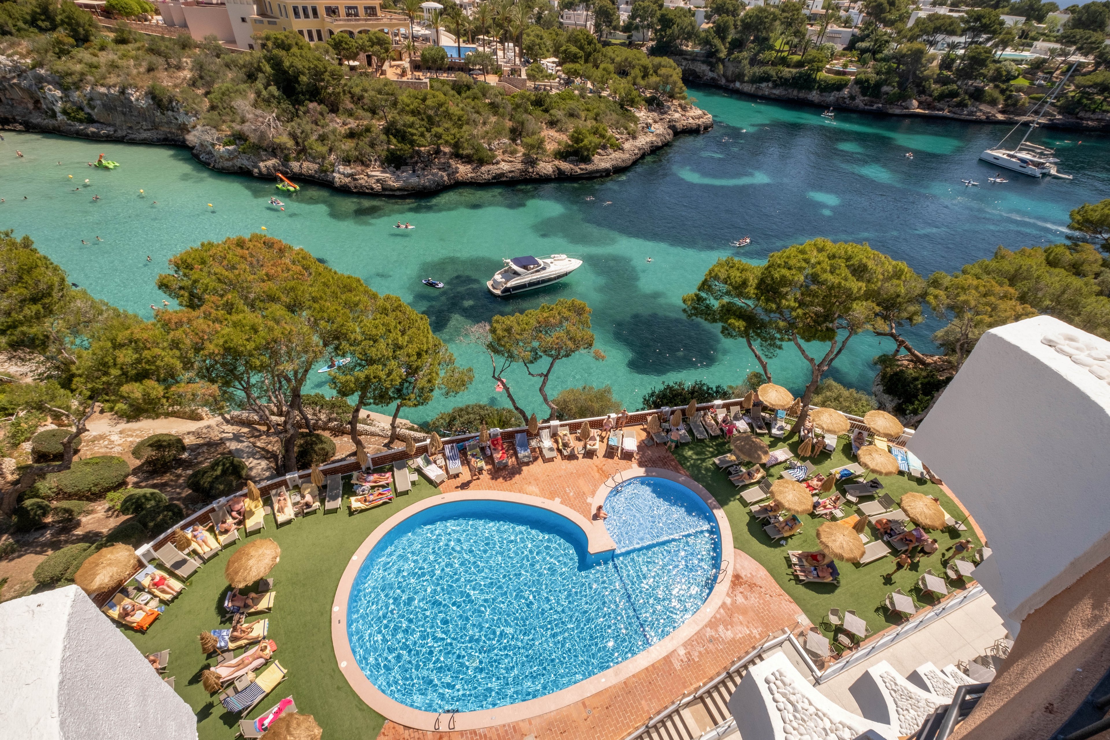 A heart-shaped outdoor swimming pool sits on a terrace above a scenic coastal inlet. Sun loungers and straw parasols surround the pool, where guests relax in the sunshine. Below, clear turquoise water fills the bay with boats and a yacht anchored near the rocky shoreline. Lush green trees and resort buildings frame the tranquil Mediterranean-style landscape.