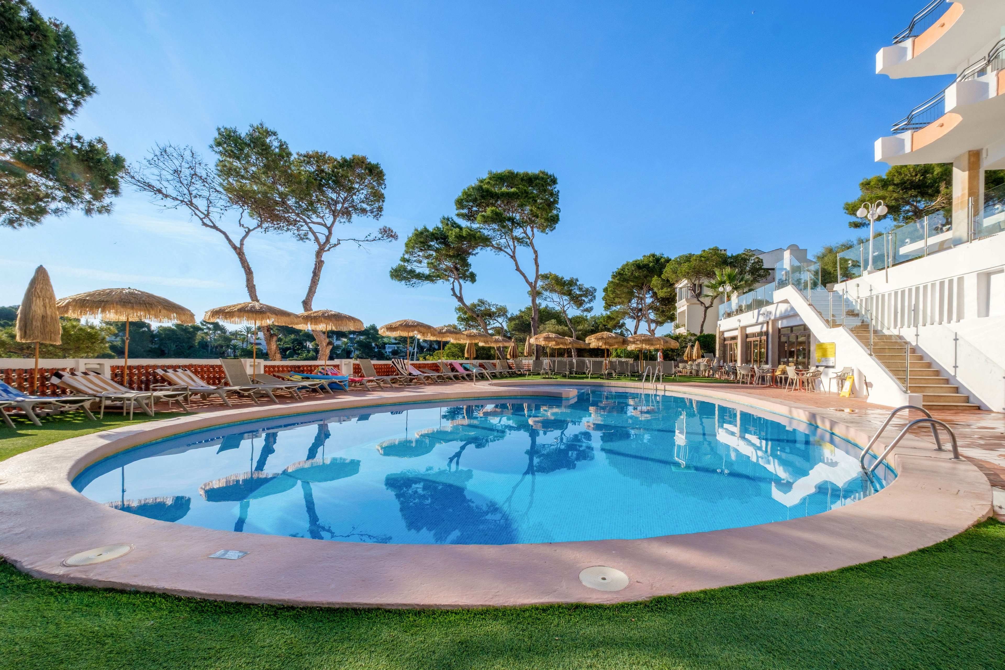 An outdoor circular swimming pool is surrounded by sun loungers and straw parasols at a resort. The scene is set on a clear, sunny day with bright blue sky and tall pine trees in the background. A modern white building with balconies and glass railings overlooks the pool area, with stairs leading down to the deck. The overall atmosphere is relaxed and inviting, ideal for vacation leisure and sunbathing.