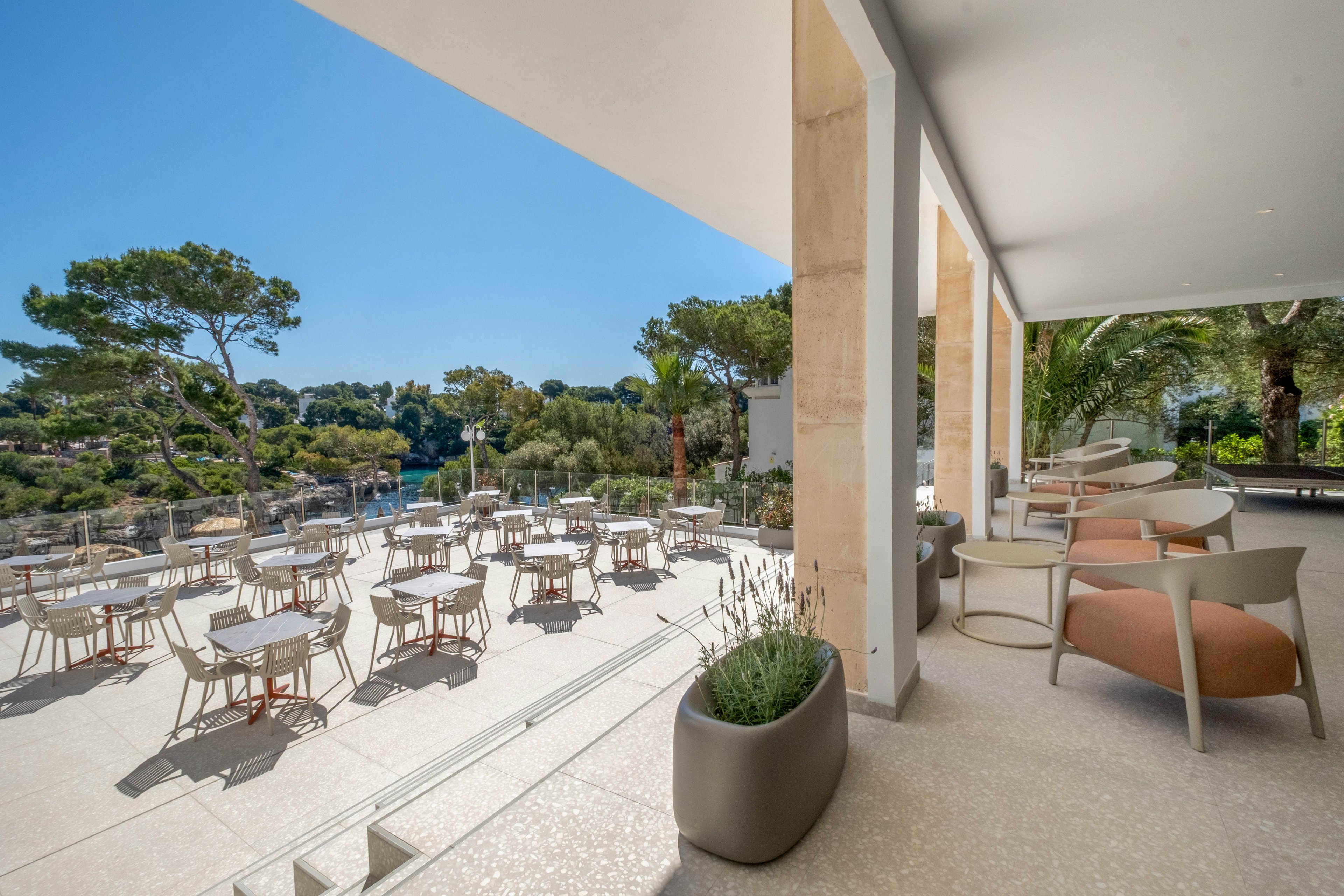 A spacious outdoor terrace features neatly arranged tables and chairs under a clear blue sky. Comfortable lounge seating lines the covered area, facing outward toward the scenic view. Beyond the terrace, lush green pine trees frame a calm waterfront, creating a serene vacation atmosphere. Large planters with ornamental plants and clean architectural lines emphasize a modern, minimalist design.