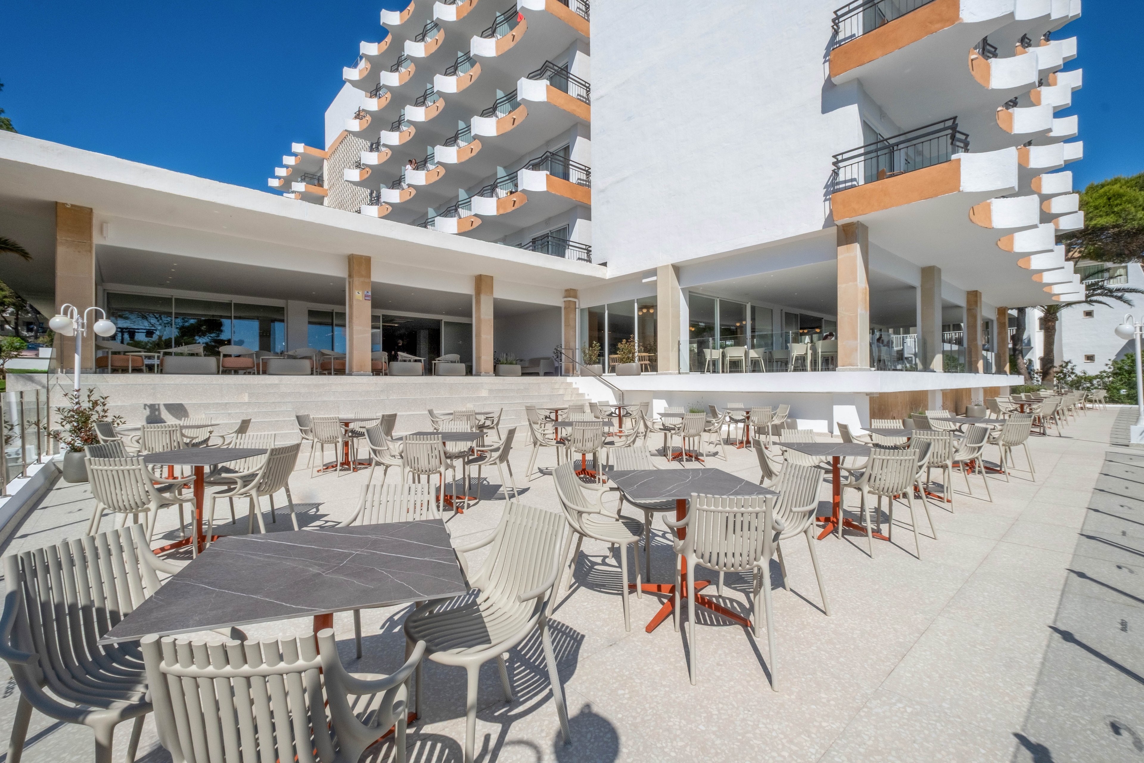 The image shows a spacious outdoor terrace with numerous tables and chairs arranged for guests. It is part of a modern white hotel building featuring multiple balconies and large glass windows. The setting appears bright and sunny, with clear blue sky and strong shadows on the ground. The overall atmosphere is relaxed and inviting, suggesting a casual dining or lounge area near a coastal destination.