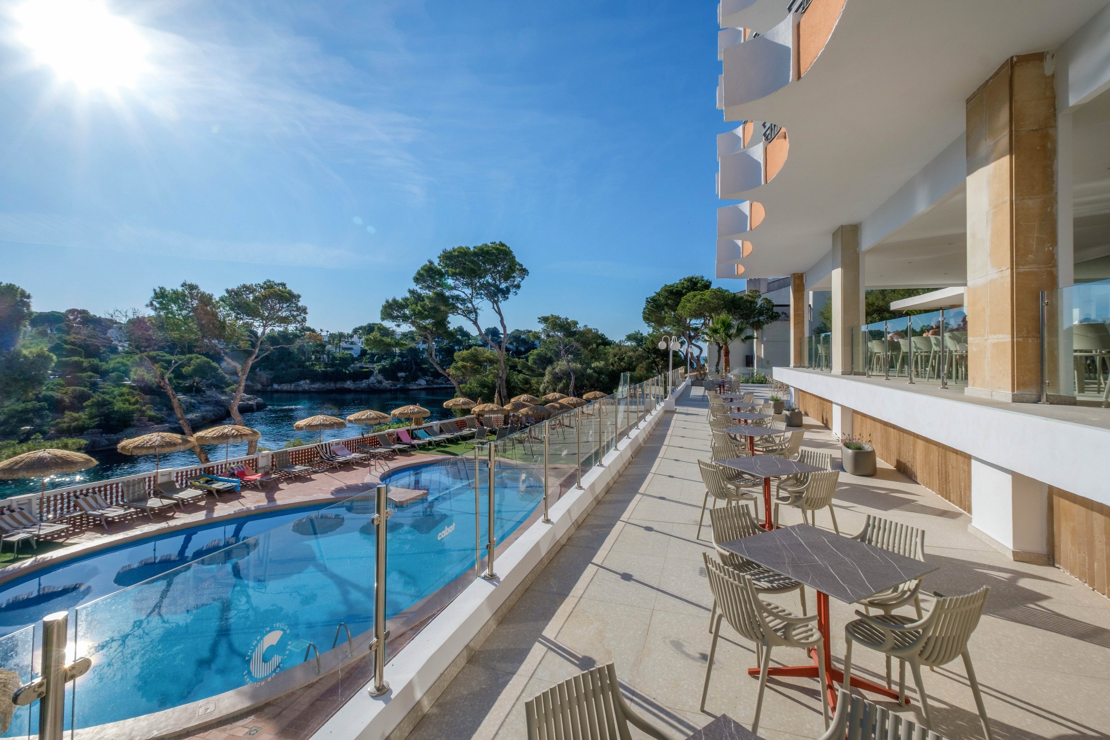 A bright resort terrace with tables and chairs overlooks a curved outdoor swimming pool. Sunlight streams across the scene, highlighting clear blue water and surrounding trees. Straw parasols line the poolside area, with a natural cove or lagoon visible beyond the railing. The atmosphere is calm and inviting, suggesting a warm vacation destination.
