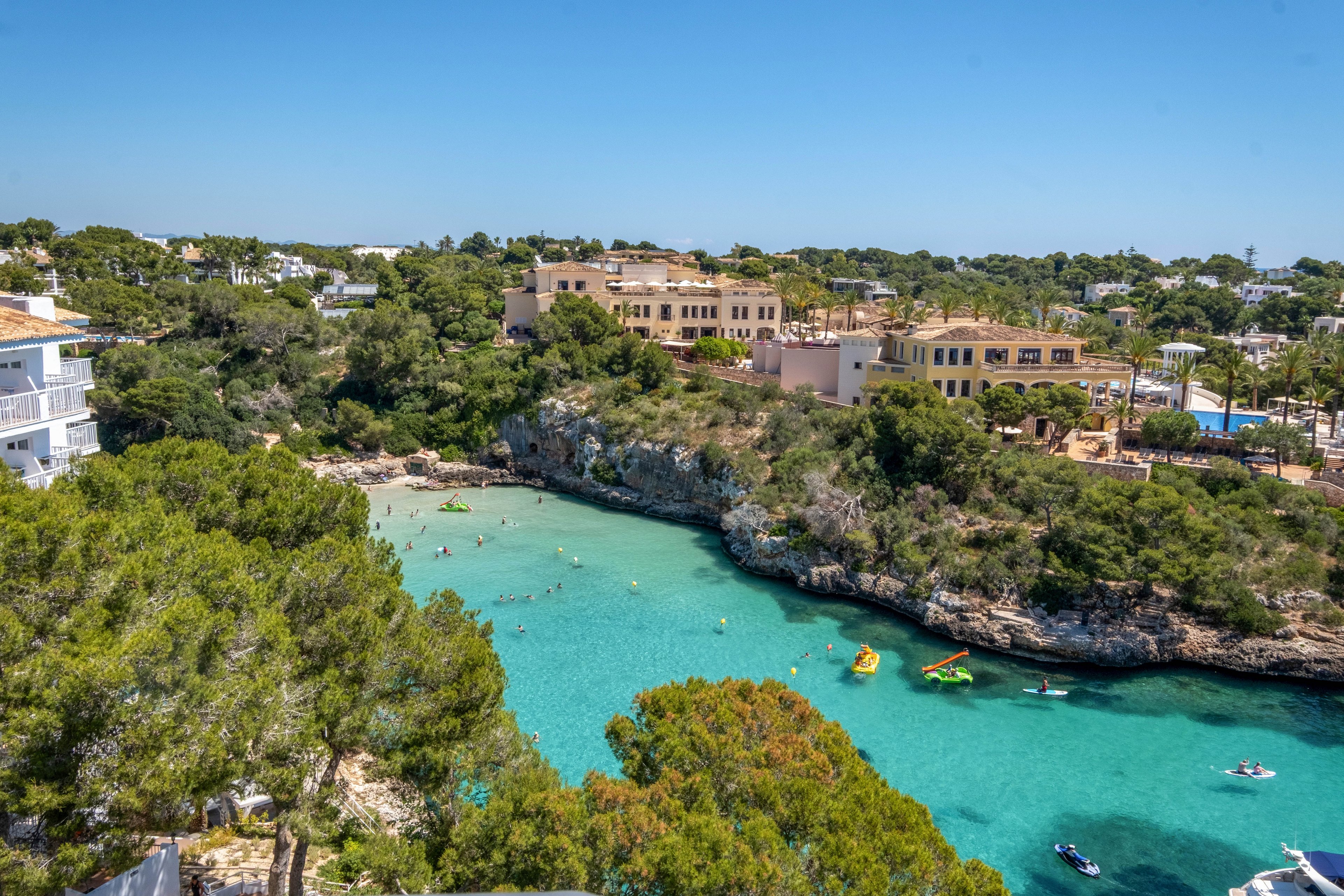 A picturesque coastal cove features bright turquoise water surrounded by rocky cliffs and lush green trees. Swimmers and kayakers enjoy the calm sea, while small boats float near the shore. Mediterranean-style buildings and villas overlook the inlet, suggesting a popular holiday destination. The clear blue sky and vibrant colors create a warm, inviting summer atmosphere.
