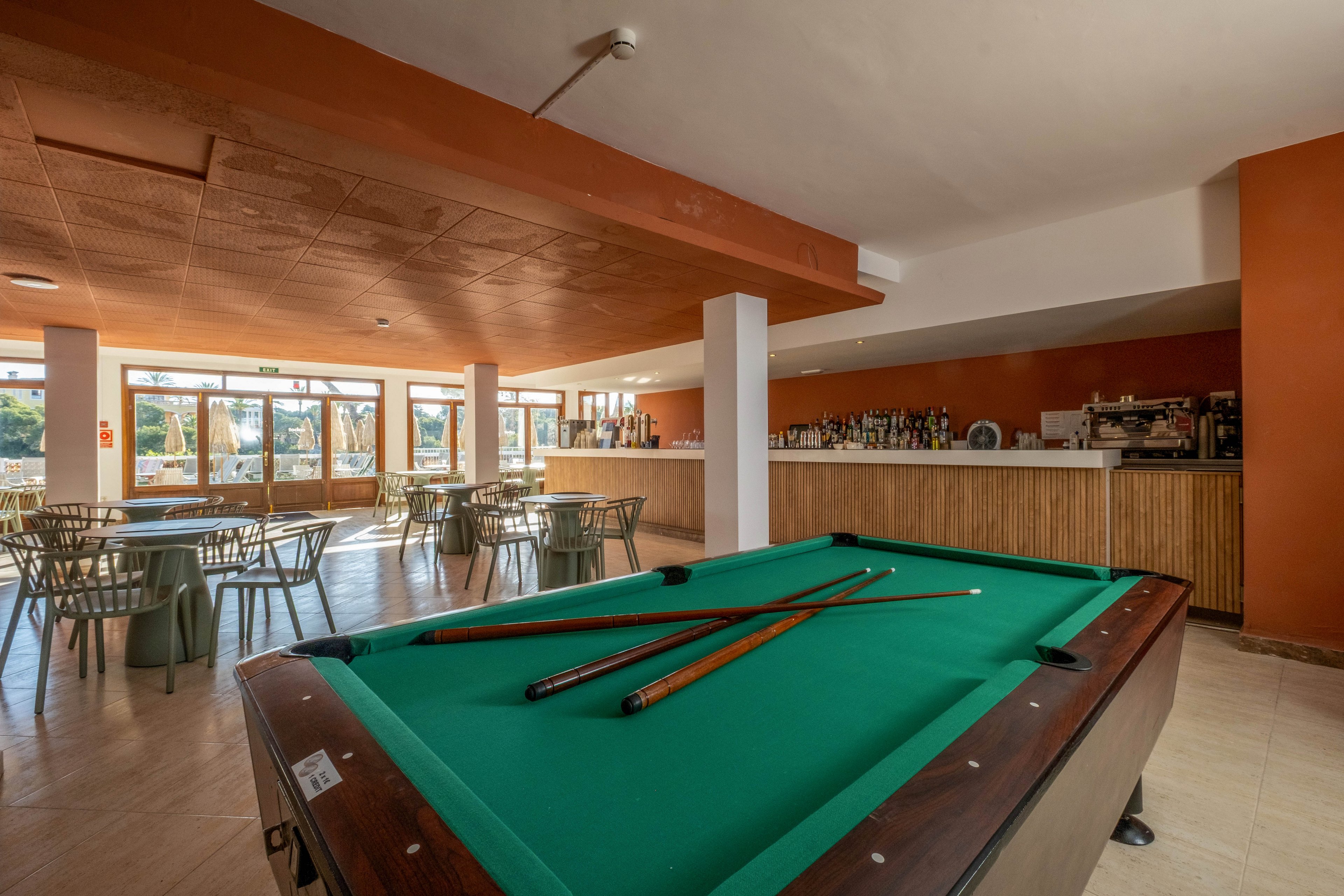 A green billiard table with cues laid across the surface stands in the foreground of a bright bar lounge. In the background, a long counter displays bottles and glassware, with bar stools and café-style tables arranged around the room. Large windows let in natural daylight, creating a relaxed and inviting atmosphere. Warm orange and neutral tones dominate the interior, suggesting a casual social space for leisure and drinks.