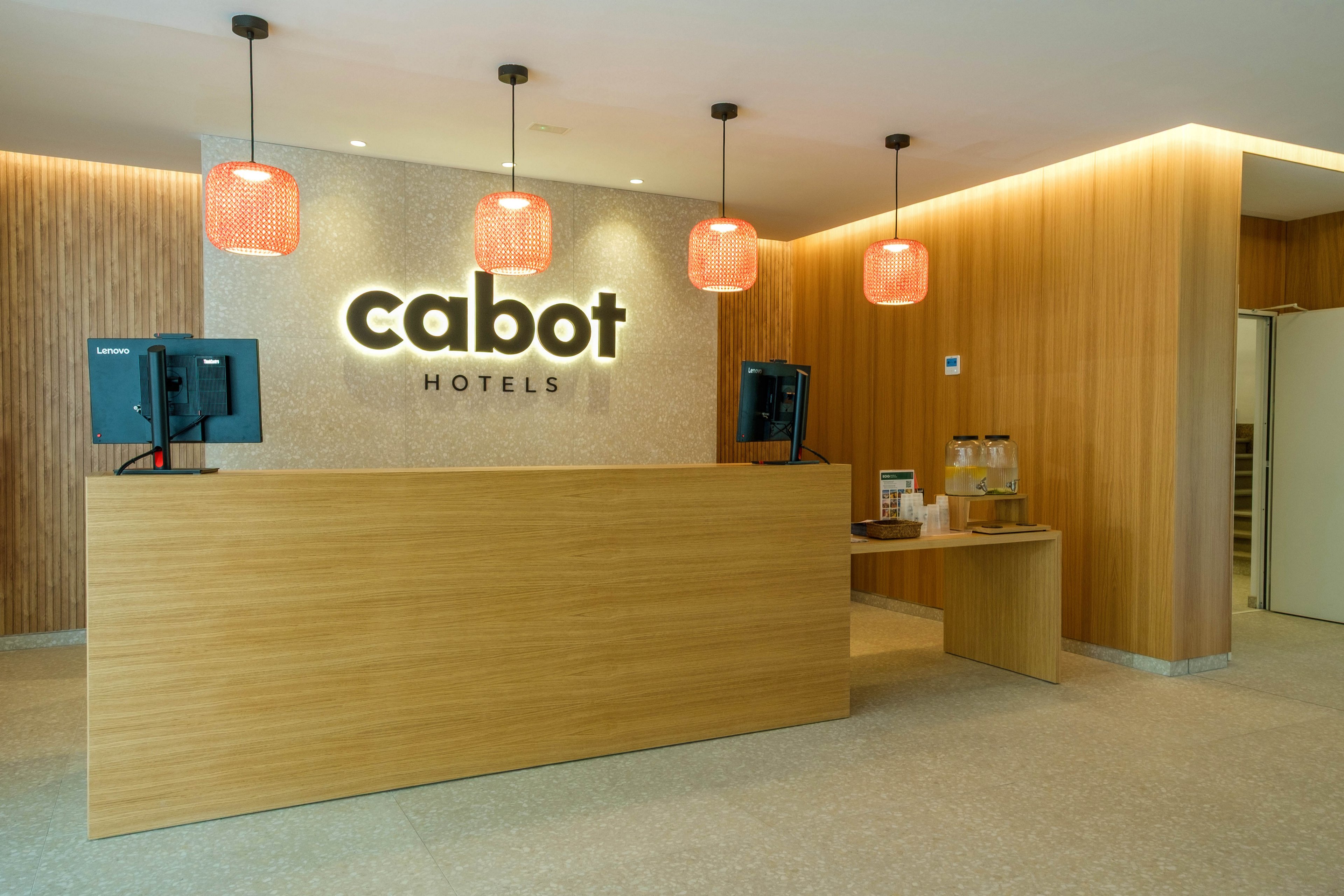 The image shows a contemporary reception desk area at Cabot Hotels. A light wood counter stands in front of a wall with illuminated Cabot Hotels signage. Warm pendant lights hang above, and the surrounding walls feature wood paneling and neutral tones. Computer monitors, beverage dispensers, and small accessories complete the welcoming hotel lobby scene.
