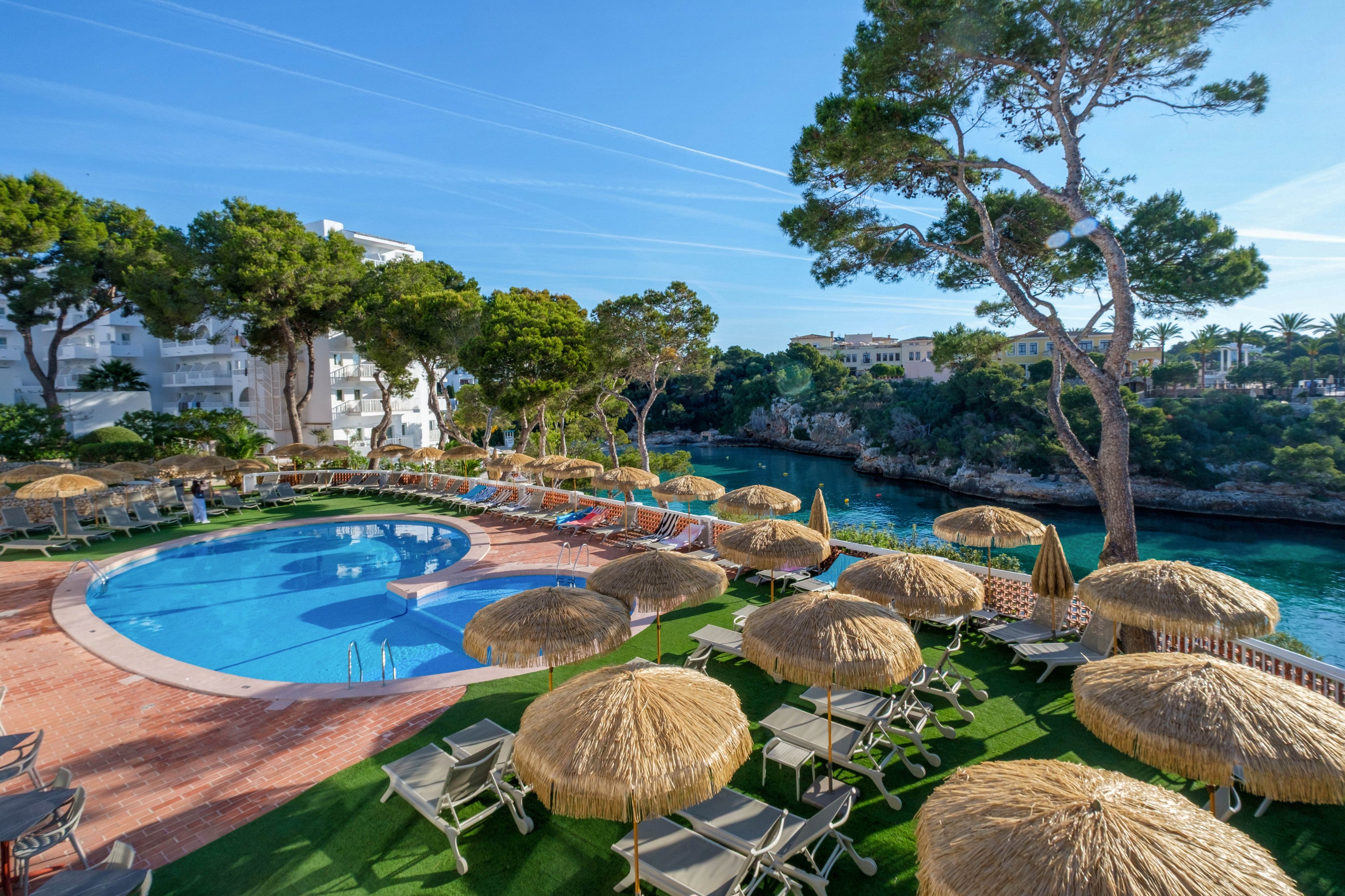 An outdoor swimming pool is surrounded by numerous straw parasols and sun loungers at a coastal resort. The setting overlooks a calm turquoise inlet bordered by rocky cliffs and lush green trees. White apartment-style buildings rise in the background under a clear blue sky with light contrails. The scene suggests a relaxing holiday atmosphere with ample seating and shade for guests.