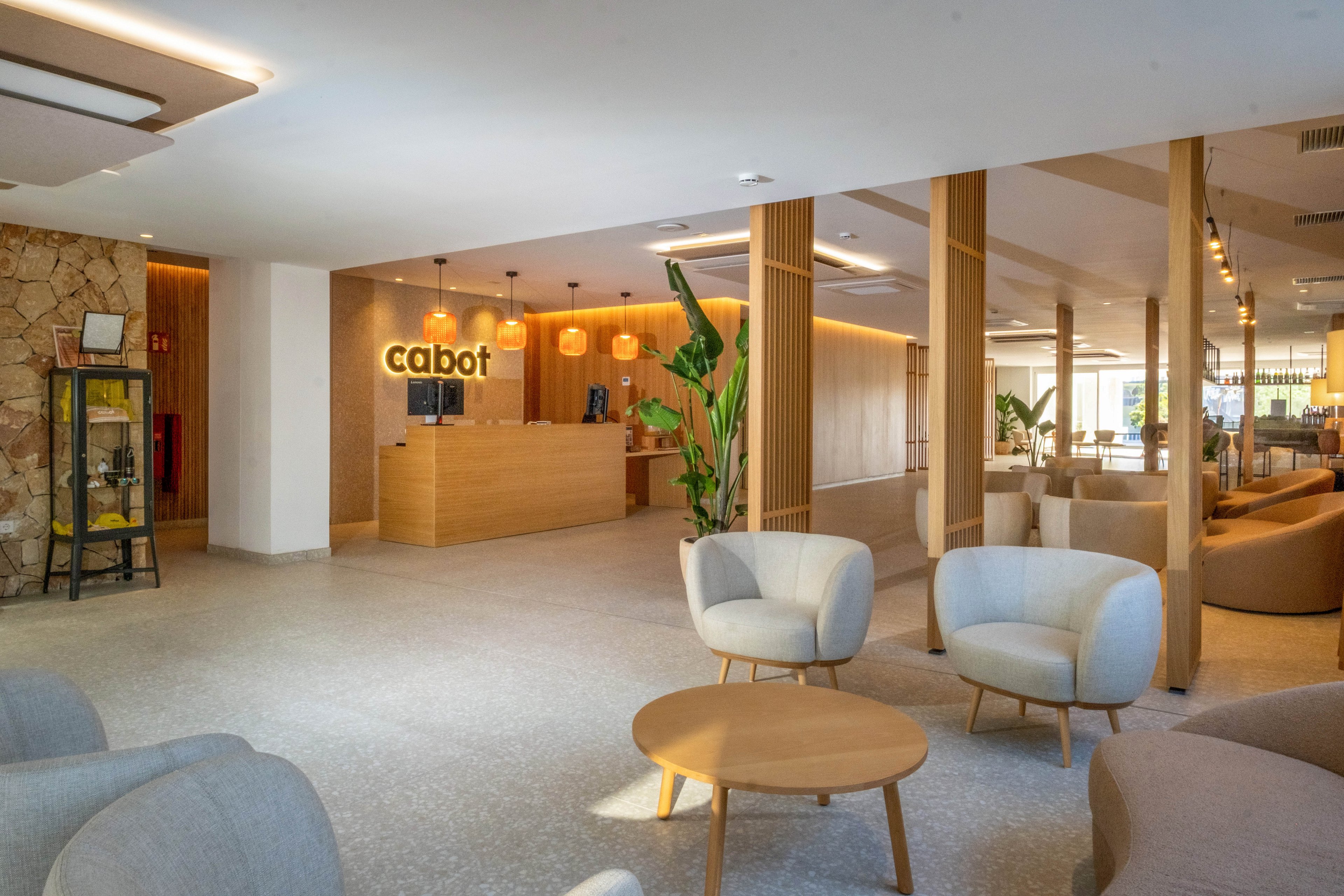 A contemporary hotel lobby features a wooden reception desk with the illuminated word cabot on the back wall. The space is open and bright, with neutral-toned armchairs and round wooden coffee tables arranged in seating areas. Vertical wooden slats, indoor plants, and warm wall lighting create a welcoming atmosphere. Stone accents and decorative shelving add texture and visual interest to the modern interior design.
