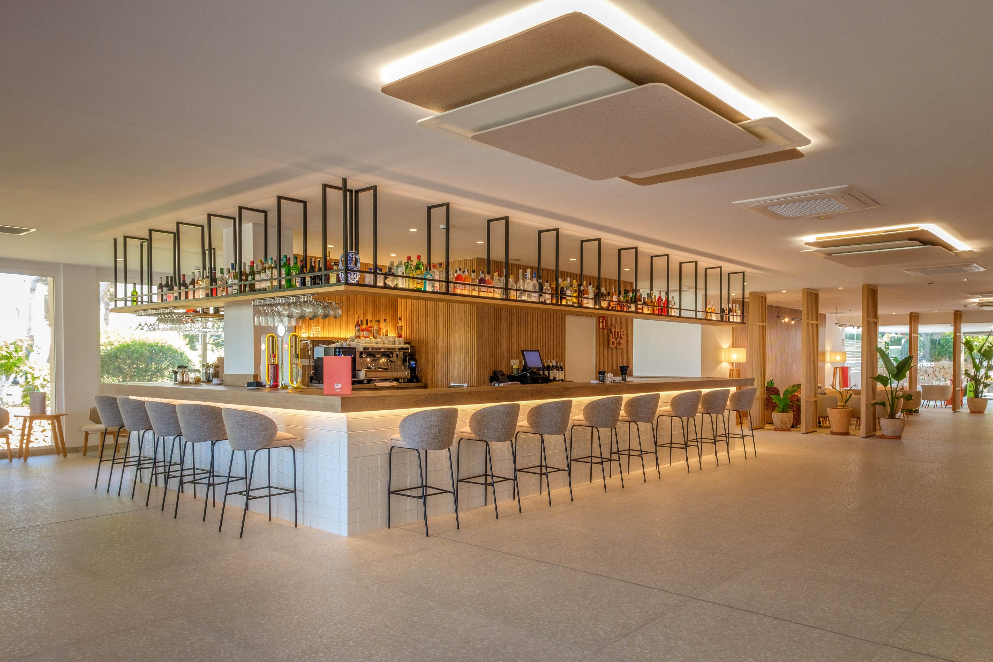 A contemporary bar counter stands at the center of a spacious, well-lit lounge. High stools line the bar, which is topped with bottles and glassware arranged on sleek shelves. Warm neutral tones, wood accents, and indirect ceiling lighting create a relaxed, upscale atmosphere. Large windows and indoor plants add brightness and a welcoming feel to the space.