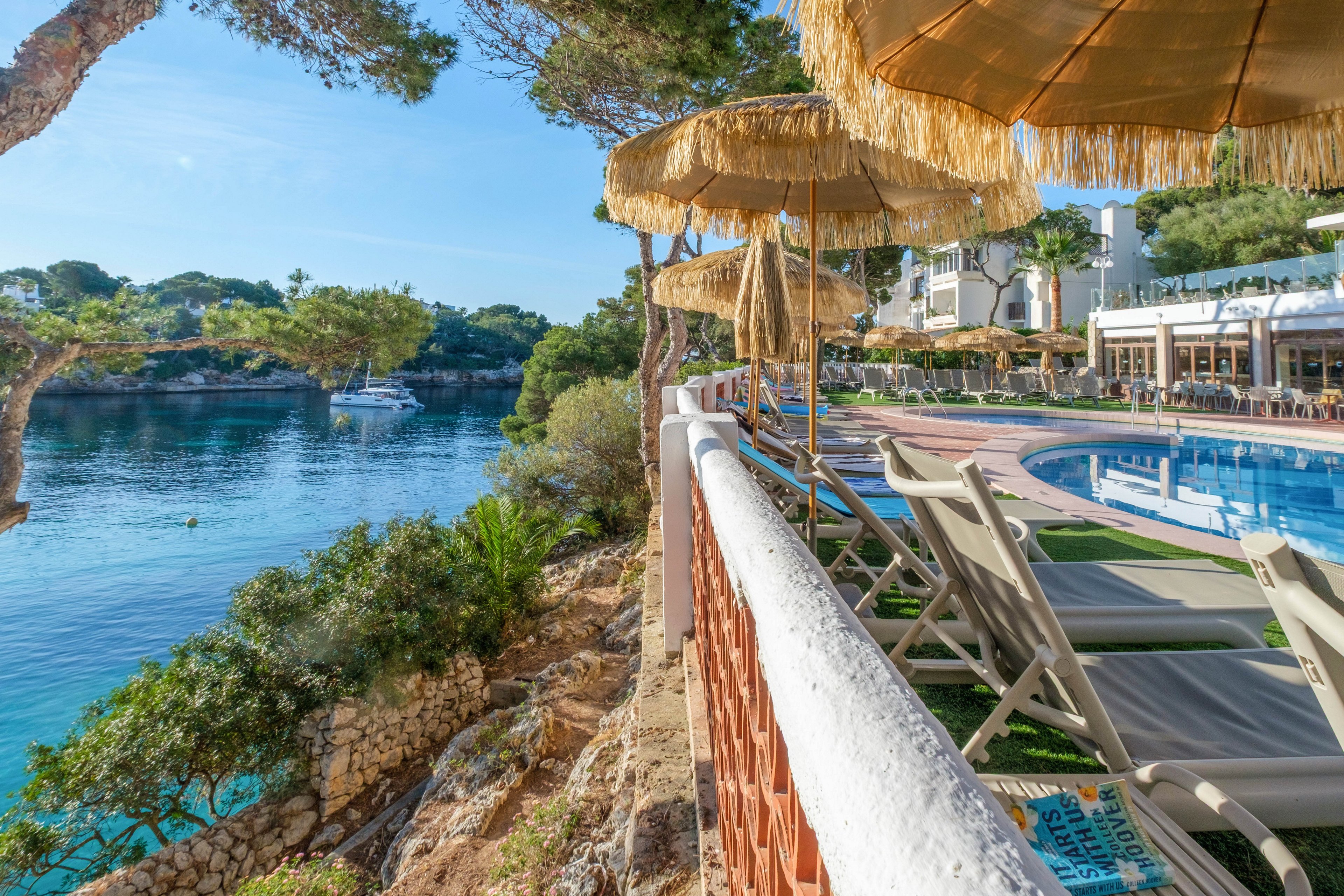A tranquil seaside resort features a row of sun loungers and straw parasols beside a clear blue swimming pool. The pool area overlooks a rocky shoreline and calm turquoise water, with trees and a boat visible in the bay. White buildings and an outdoor terrace suggest a vacation hotel setting under a bright, sunny sky. The overall scene conveys relaxation and leisure in a coastal holiday destination.
