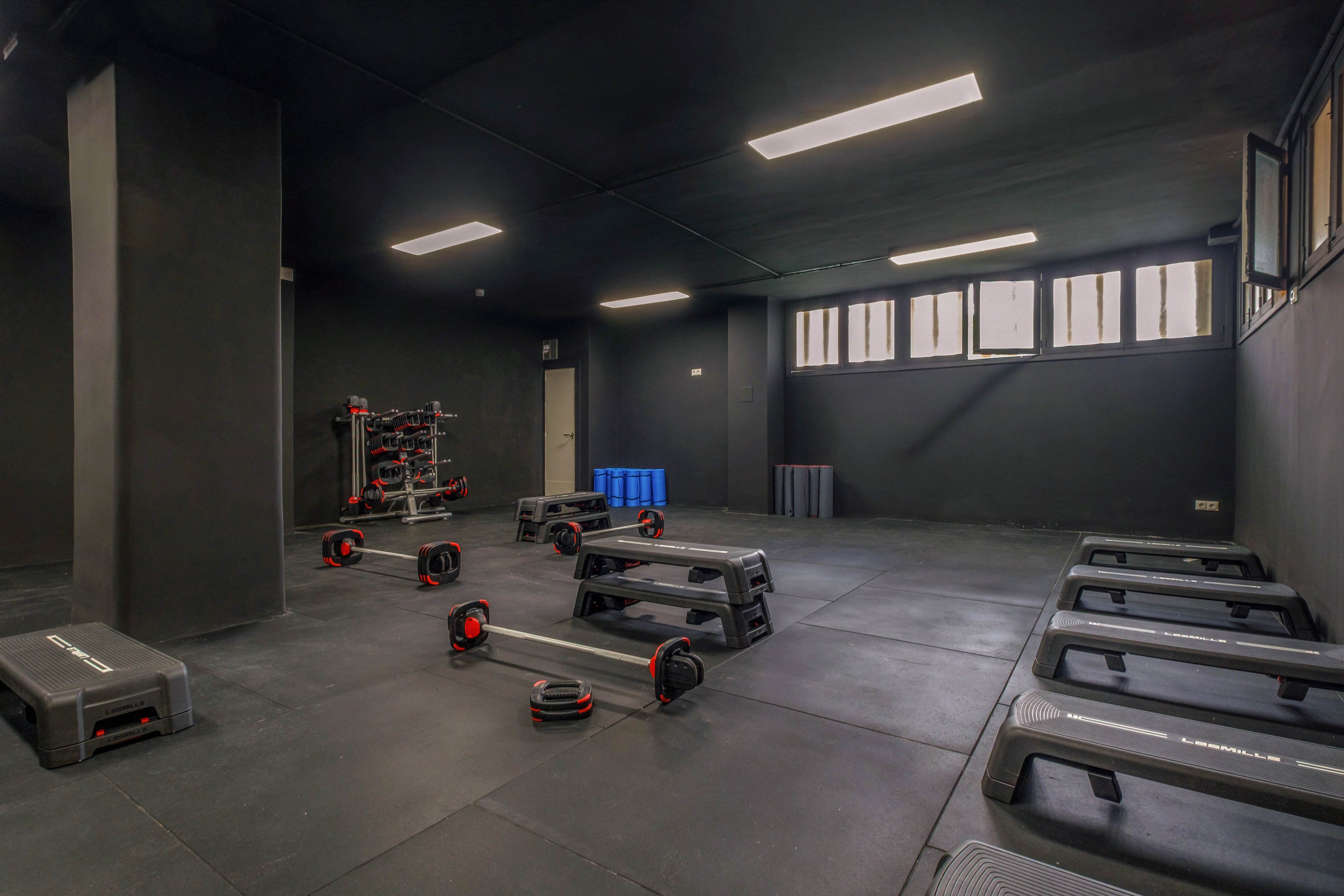 A spacious, empty fitness studio features multiple step platforms arranged on a dark rubber floor. Weight bars and plates are stored on a rack in the corner, with additional equipment neatly organized nearby. Overhead fluorescent lights illuminate the minimalist, industrial-style room with dark walls and ceiling. High windows along one side bring in natural light, emphasizing the clean and orderly workout space.