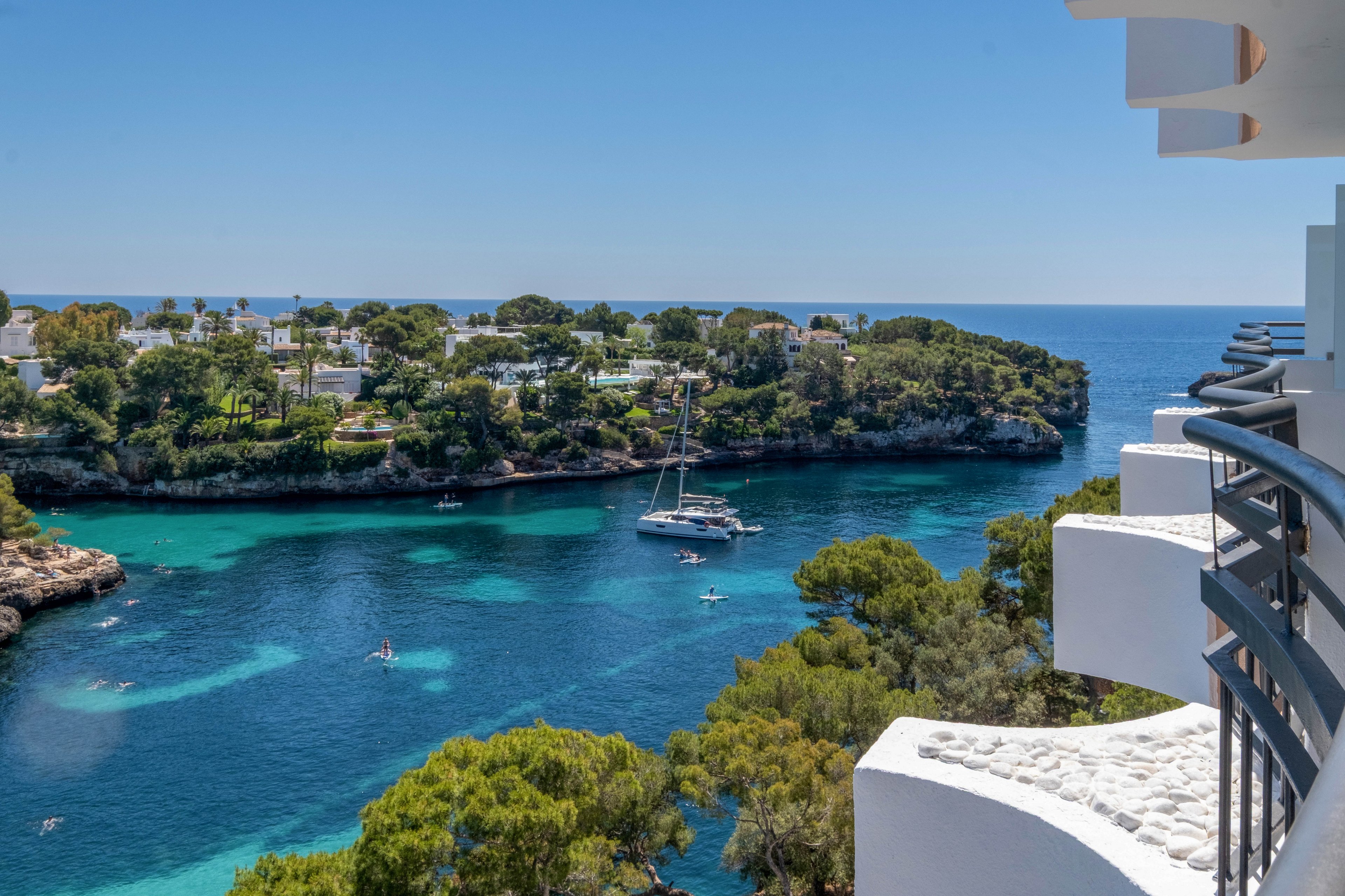 A scenic view from a white hotel balcony overlooks a calm turquoise cove. A sailboat and several small boats float on the clear blue water surrounded by lush green trees. White buildings and villas line the opposite shore under a bright blue sky. The image conveys a sunny, relaxing coastal holiday atmosphere.