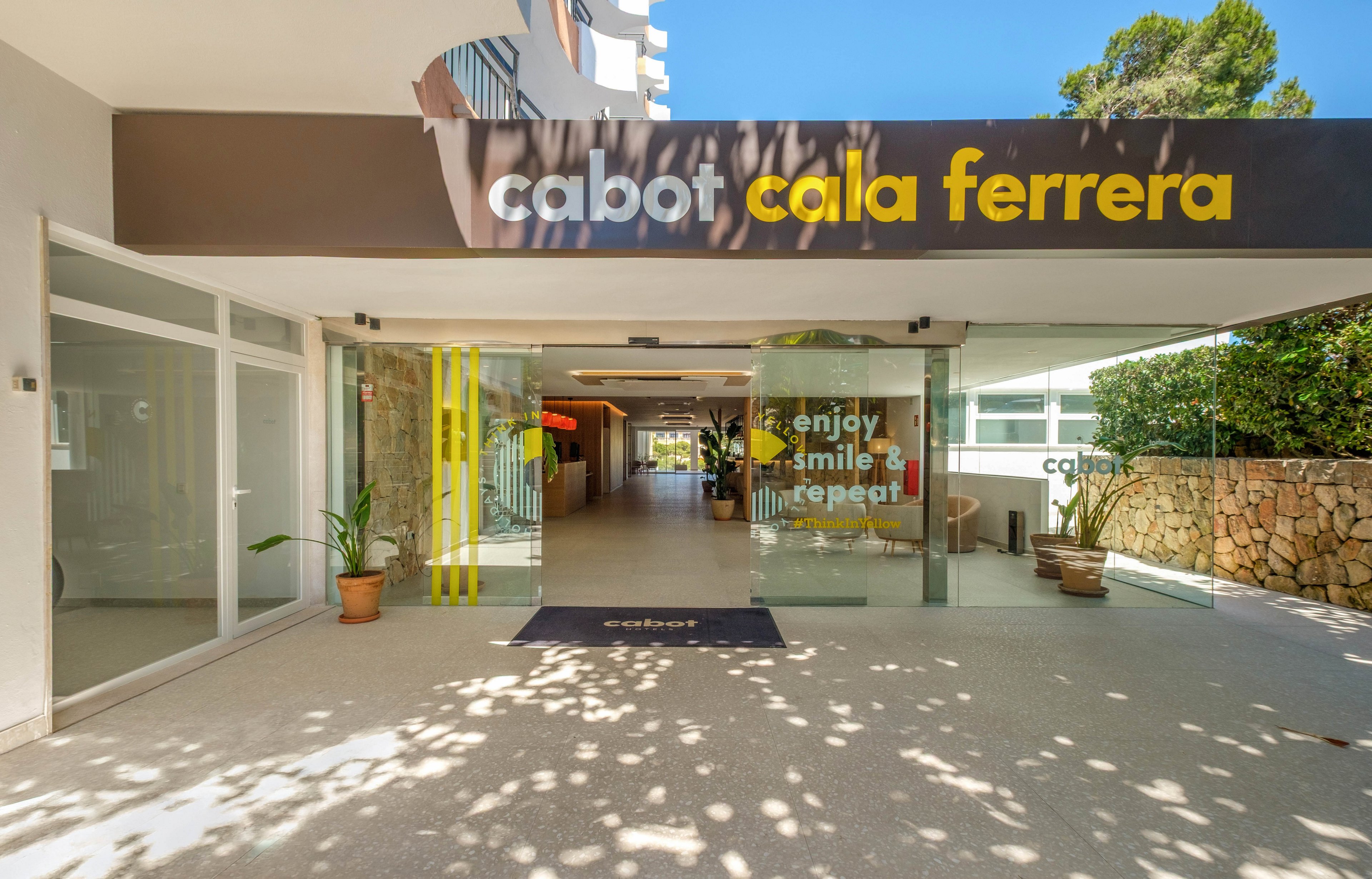 Wide view of the Cabot Cala Ferrera hotel entrance with a covered walkway leading inside. Large signage with the name is displayed above the entry, and glass doors reveal a bright interior corridor. A welcome mat and potted plants frame the doorway, while a stone wall and greenery line the side. Text on the glass inside reads "enjoy smile & repeat," adding a cheerful, inviting feel to the scene.