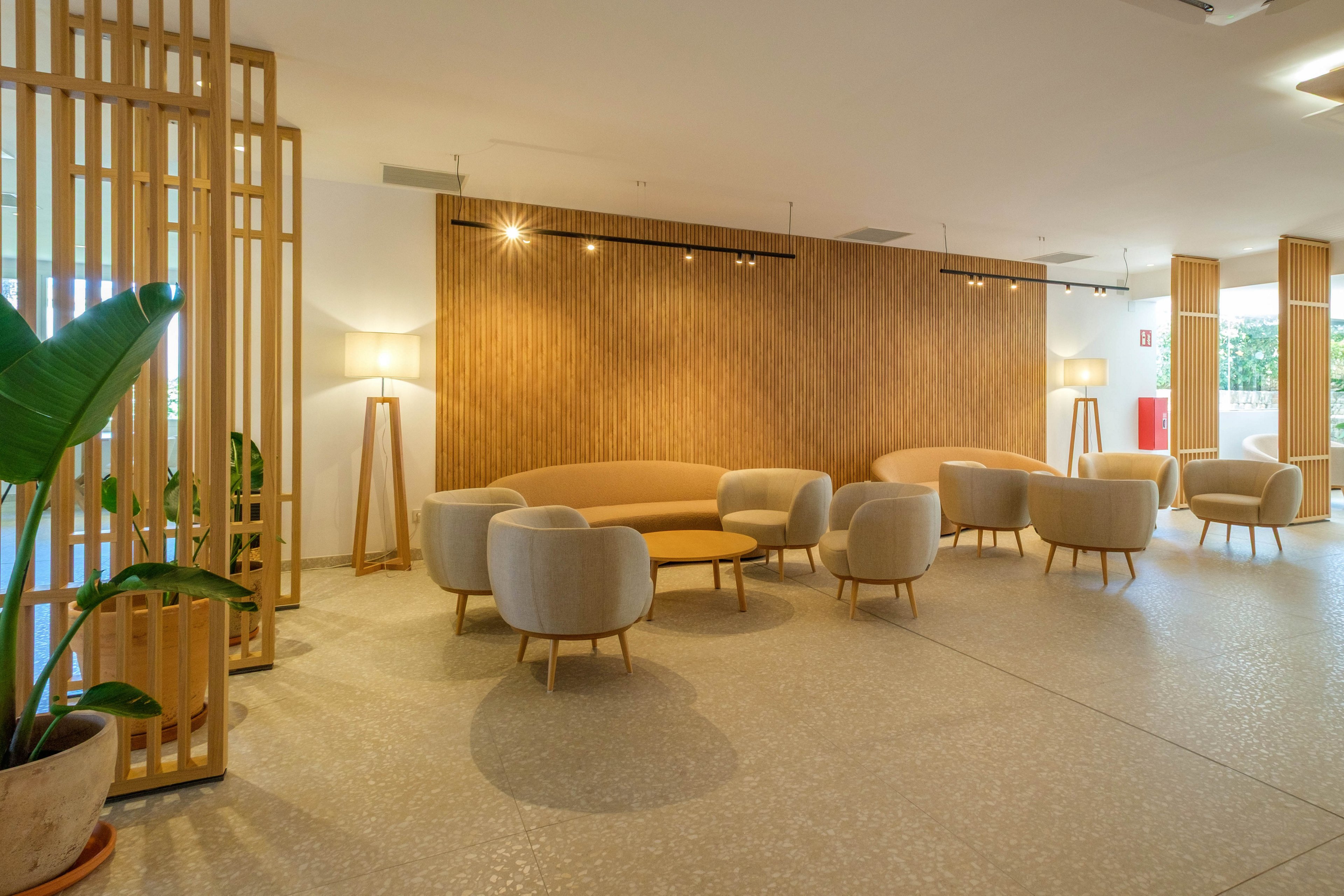 A contemporary lounge features curved sofas and cushioned armchairs arranged around low round tables. The space is defined by a warm wooden slat wall and matching vertical room dividers. Soft ambient lighting from floor lamps and ceiling spotlights creates a calm, inviting atmosphere. A large potted plant adds a touch of greenery to the minimalist interior design.