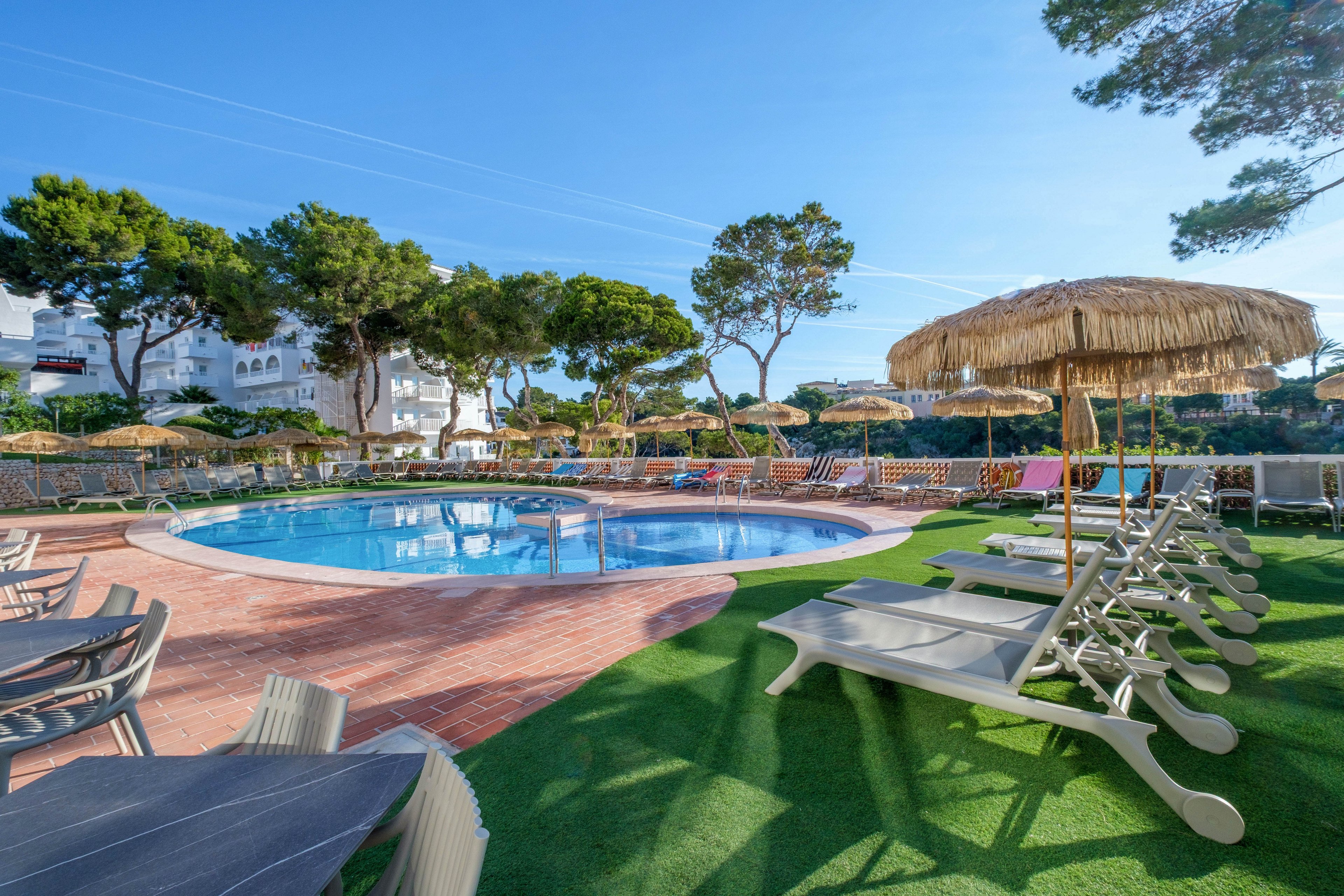 An outdoor resort swimming pool is surrounded by sun loungers and straw parasols under a clear blue sky. The area features artificial grass and red brick paving, creating a neat and inviting relaxation space. White buildings and tall green trees frame the poolside, suggesting a holiday destination near nature. The scene is brightly lit, emphasizing a warm, sunny atmosphere ideal for vacation leisure.
