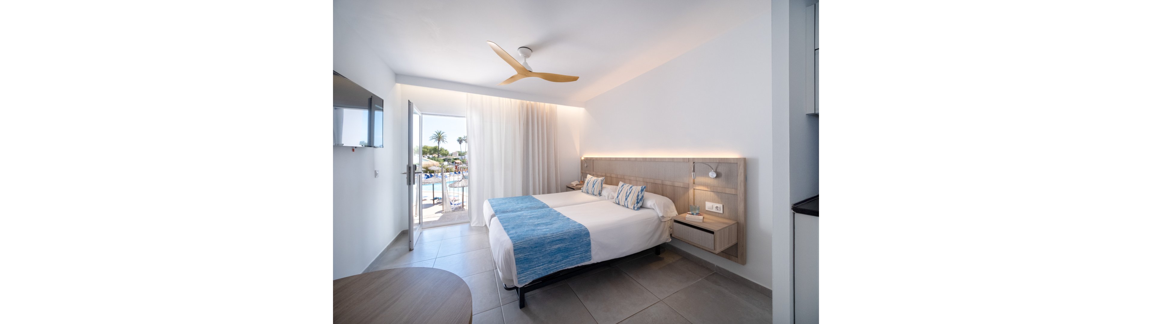 A modern hotel room features a neatly made double bed with white linens and blue accents. The space is bright and minimal, with light gray floor tiles and a wooden headboard with integrated lighting. An open door leads to a balcony overlooking a sunny pool area with palm trees. A ceiling fan, wall-mounted TV, and compact bedside tables complete the contemporary vacation setting.