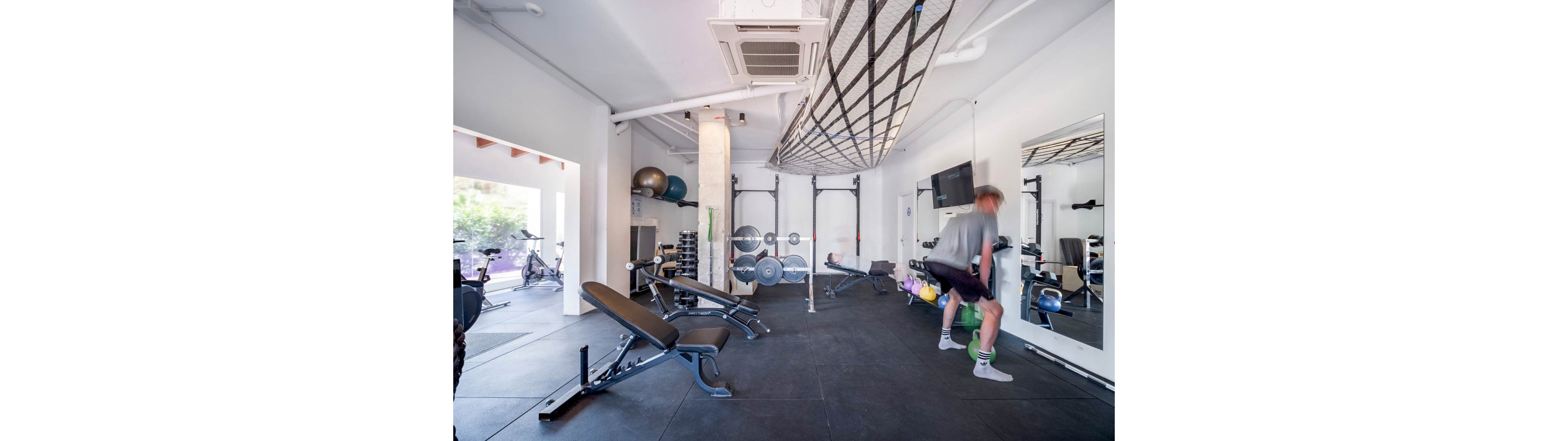 A man exercises with a weight in a bright, modern gym. The space features strength training equipment, benches, medicine balls, and a large functional rig. A rope net is suspended from the ceiling and a bicycle is visible near the open entrance. The overall atmosphere is clean, spacious, and focused on fitness and performance.