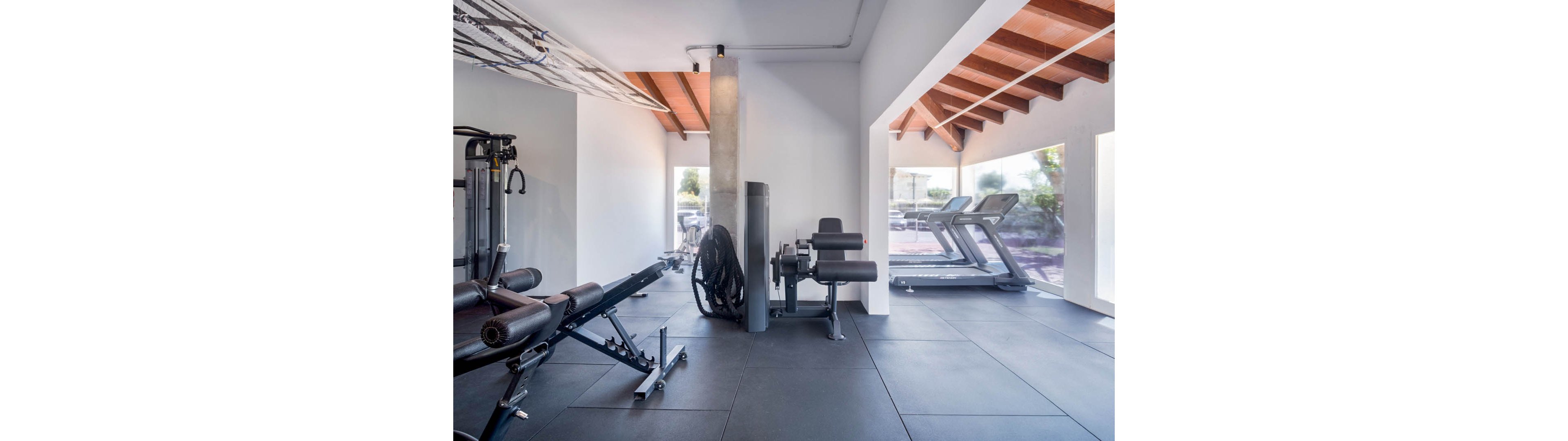 A bright, modern gym room features various strength and cardio machines. Weight benches and resistance equipment are arranged on black rubber flooring in the foreground. In the background, two treadmills face large windows that let in natural light and views of the outdoors. Exposed wooden ceiling beams and white walls create a clean, contemporary fitness environment.