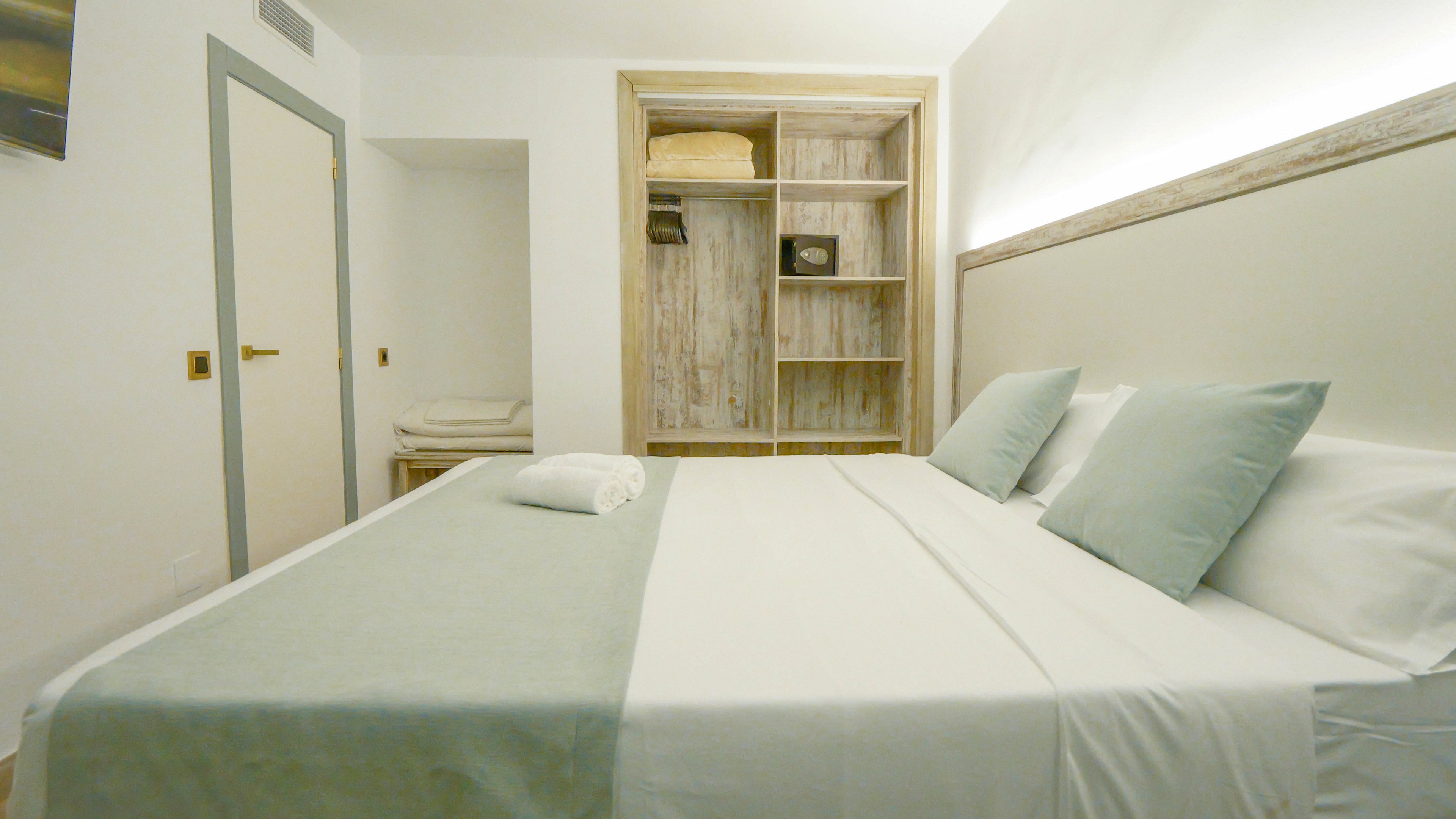 A neatly made double bed with pale green accents dominates a minimalist bedroom. The room features a built-in open wardrobe with folded linens and shelves, and a closed door on the left side. Soft, indirect lighting along the headboard creates a calm and inviting atmosphere. Neutral colors and clean lines emphasize a modern, uncluttered design.