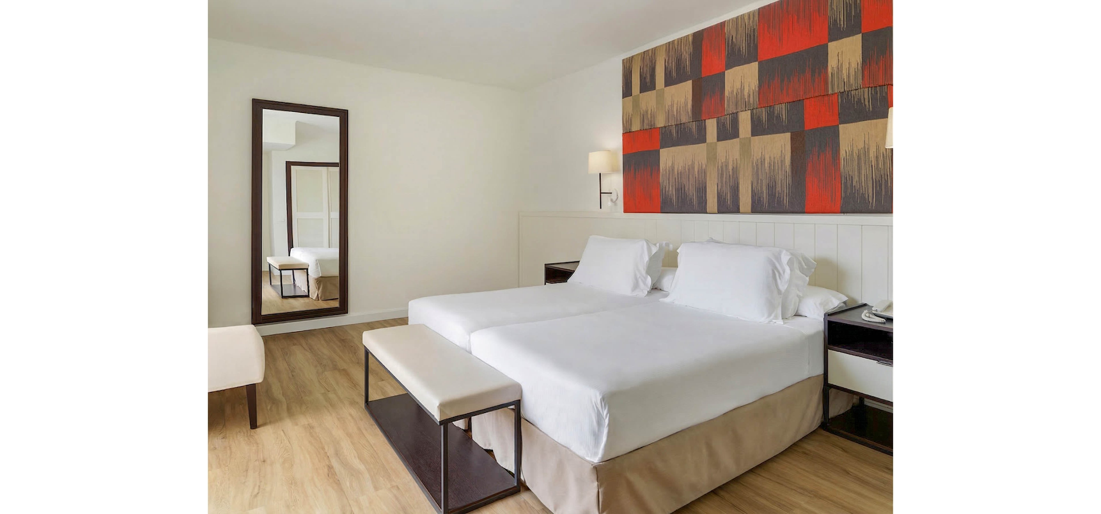 A contemporary hotel room features two neatly made twin beds with white linens and a shared upholstered bench at the foot. The space has light wood flooring, a large wall mirror, and a simple bedside table with a phone. Warm wall-mounted lamps and a bold geometric artwork above the beds add color and visual interest. The overall design is clean, minimal, and inviting, suggesting a comfortable stay for guests.