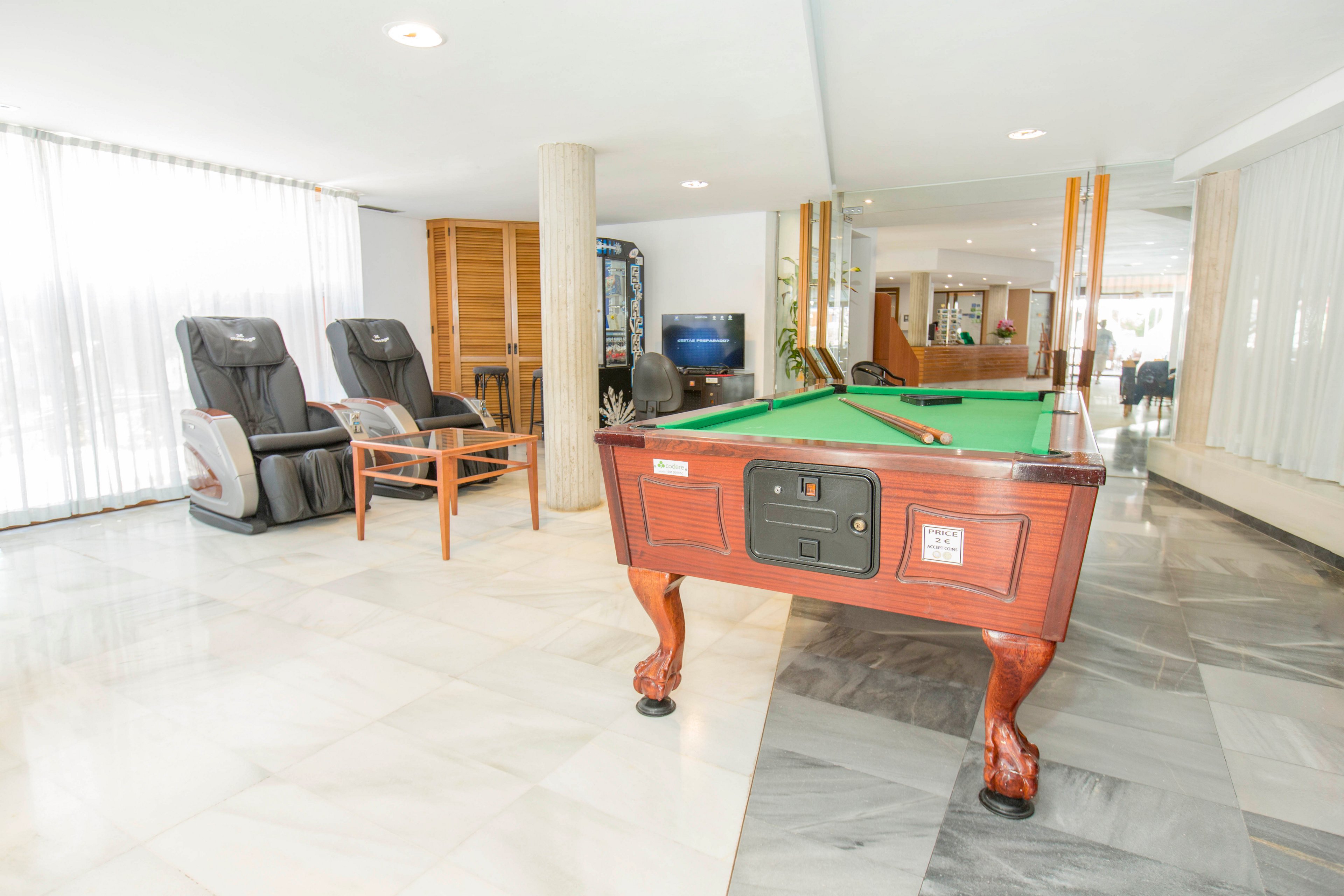 A wooden pool table with green felt stands prominently in a bright recreation lounge. Nearby, two massage chairs and a small wooden table with chairs create a relaxed seating area. Large windows with sheer curtains let in abundant natural light, reflecting off the polished marble floor. In the background, additional furniture and entertainment machines contribute to a casual, leisure-focused atmosphere.