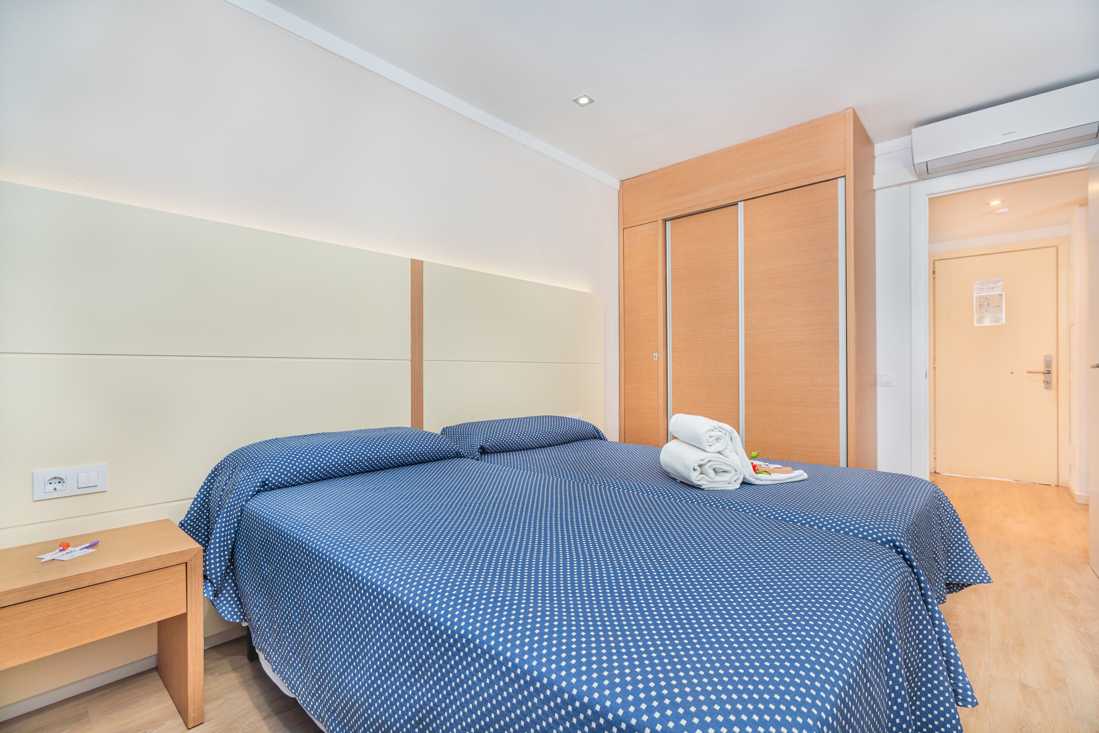 A clean, modern hotel room features two twin beds pushed together, covered with blue polka dot bedspreads. The space is bright with light-colored walls, wooden furniture, and a large built-in wardrobe. Fresh white towels are neatly folded on the bed beside a small tray. A bedside table with a keycard and an air conditioning unit above the door complete the simple, functional design.