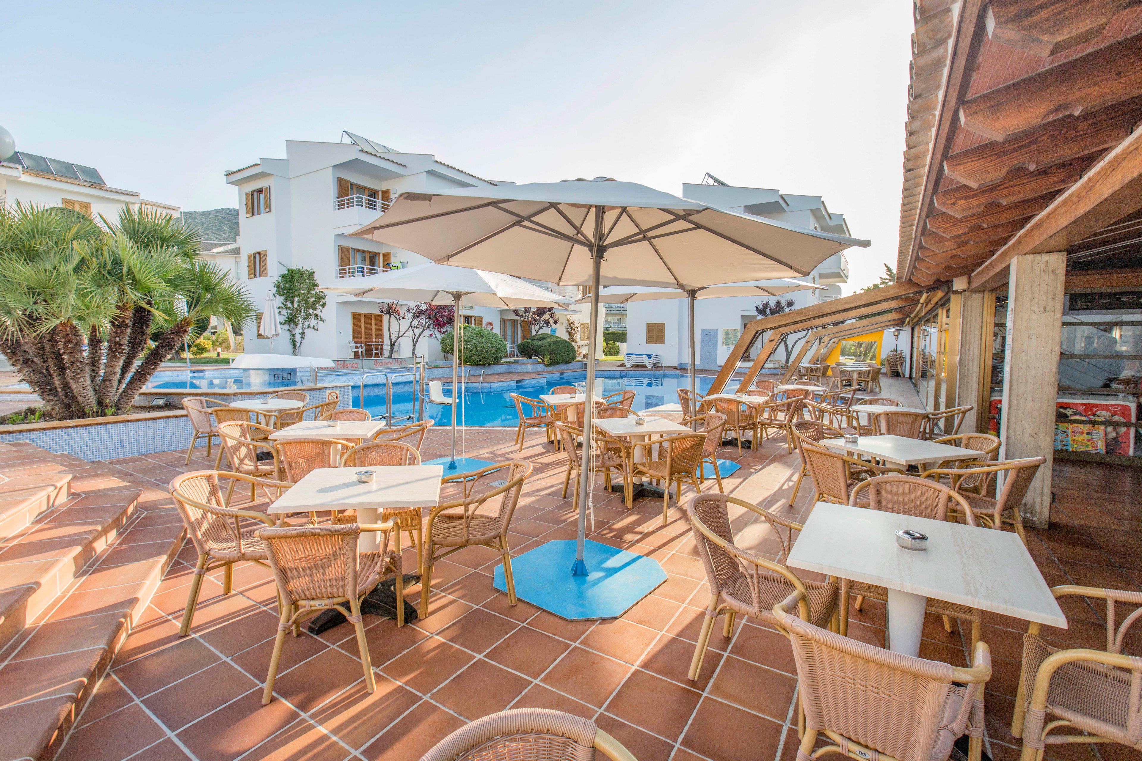 An outdoor resort terrace cafe overlooks a bright blue swimming pool. Wicker chairs and light wooden tables are arranged under large white umbrellas on a terracotta-tiled patio. A modern white hotel building with balconies stands in the background, framed by palm trees and clear sky. The scene feels relaxed and inviting, suggesting a warm vacation setting.