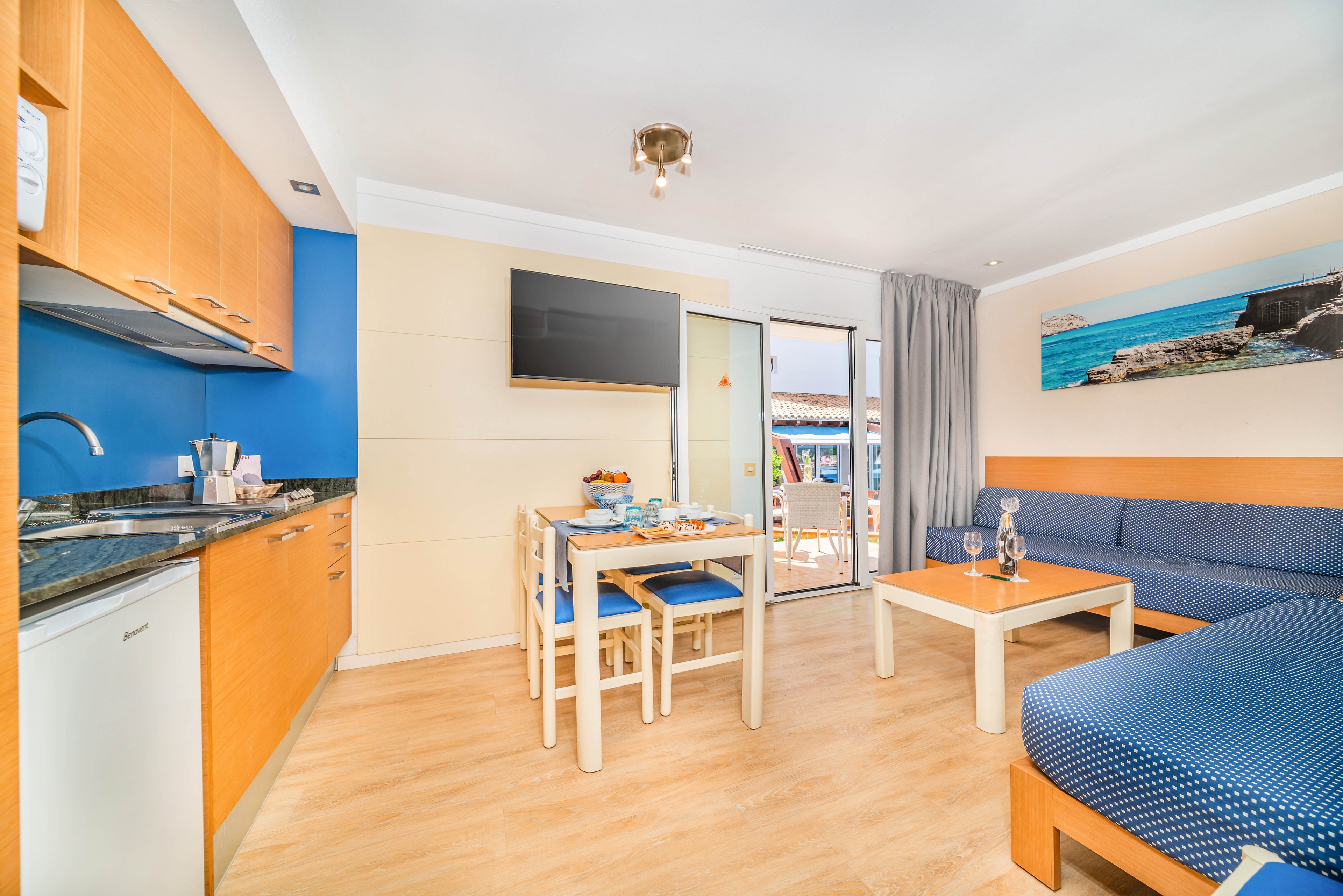 A modern studio apartment features a compact kitchenette and dining area with light wood cabinetry and blue accent walls. A flat-screen television is mounted above the dining table, which is set for a meal. A cozy seating area with blue upholstered sofas and a coffee table sits near a large sliding glass door leading to an outdoor terrace. Natural light fills the room, highlighting the clean, contemporary decor and coastal artwork on the wall.