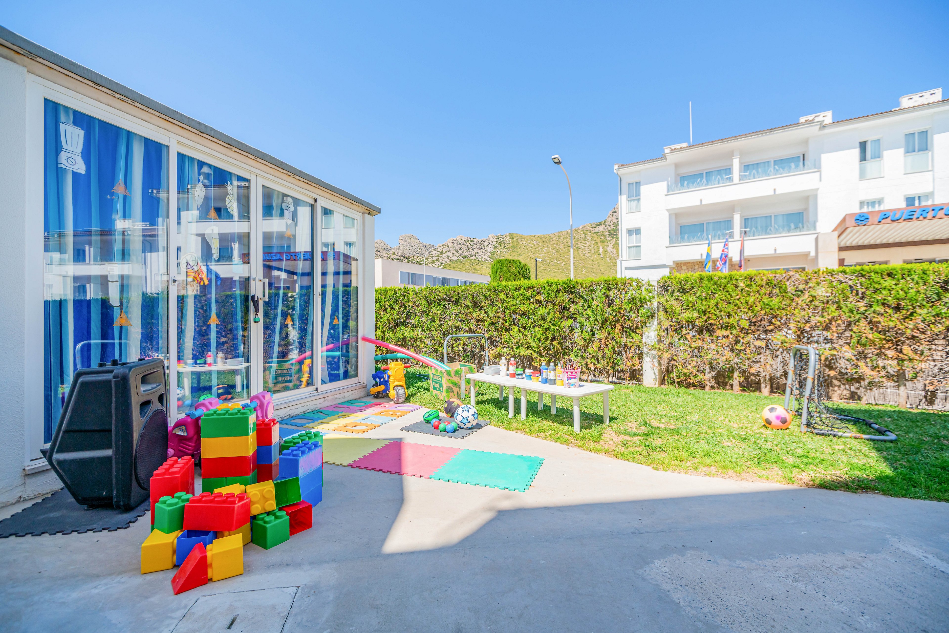 A bright outdoor children's play area is set up beside a glass-walled building at a resort. Large foam blocks, mats, and various toys are arranged on the ground and on small tables. A trimmed hedge separates the play zone from nearby white buildings under a clear blue sky. The scene suggests a safe, sunny space for kids to play and socialize.