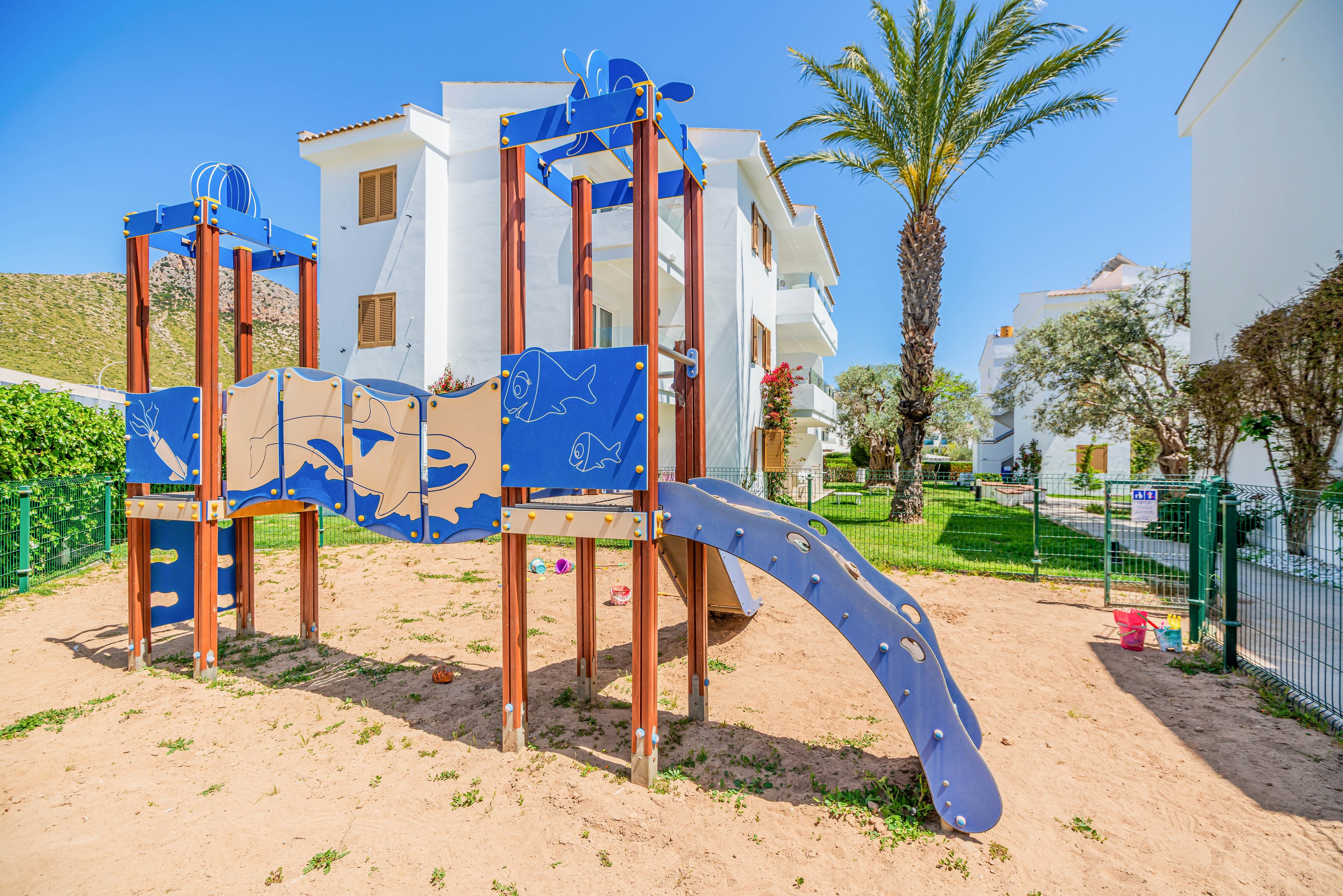 A modern playground structure with blue panels and a curved slide stands on a sandy surface in a residential courtyard. The play equipment features marine-themed illustrations such as fish and waves. White apartment buildings and balconies surround the play area, with a tall palm tree adding a tropical touch. Bright sunlight and a clear blue sky create a cheerful, family-friendly atmosphere.