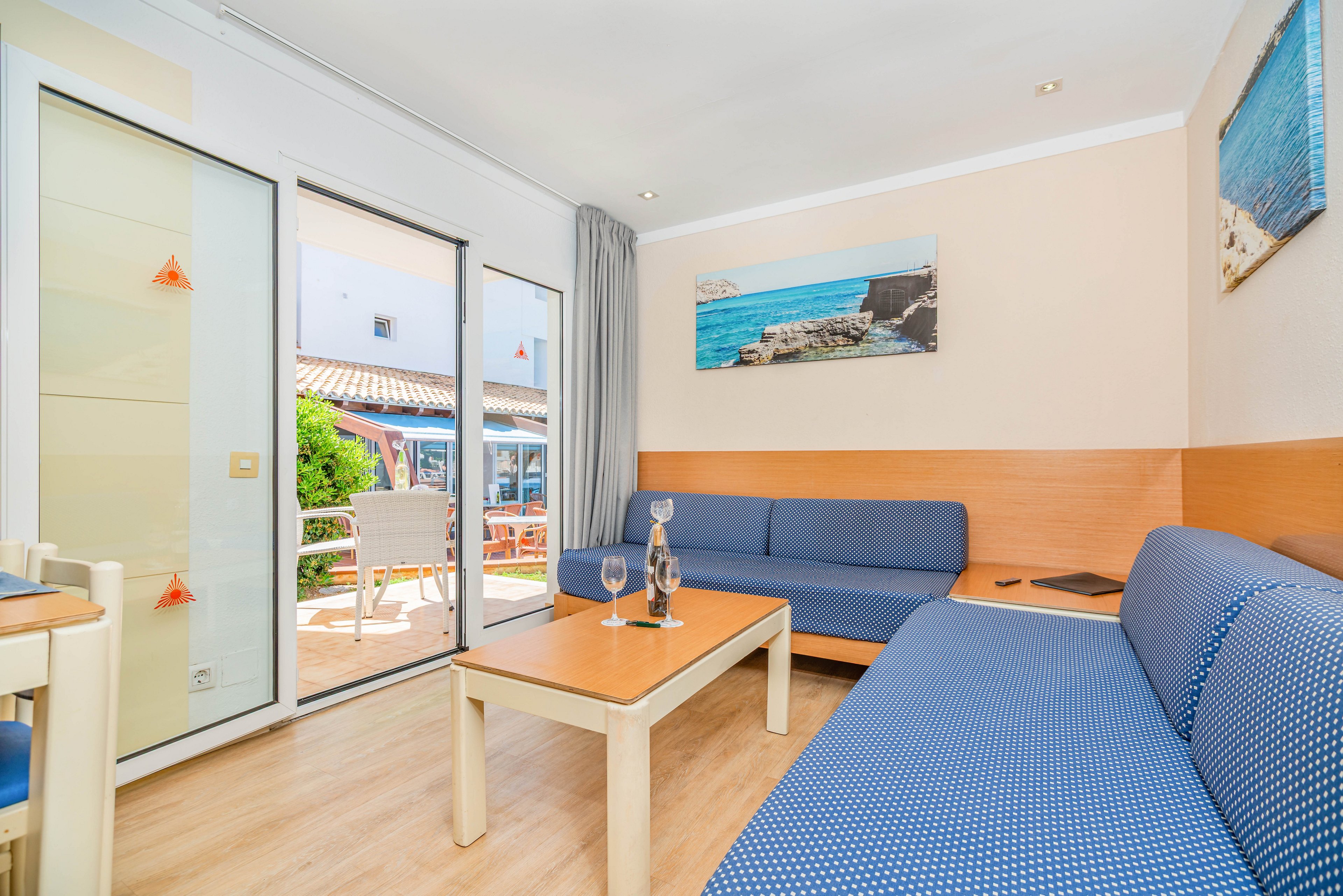 A modern hotel suite features blue upholstered sofas and a light wooden coffee table with a glass bottle and wine glasses. Large glass doors open onto a sunny terrace with outdoor chairs and a view of neighboring buildings. Wall art depicting a coastal scene and neutral-colored walls enhance the seaside ambiance. Natural light fills the room, creating an inviting and relaxed atmosphere.