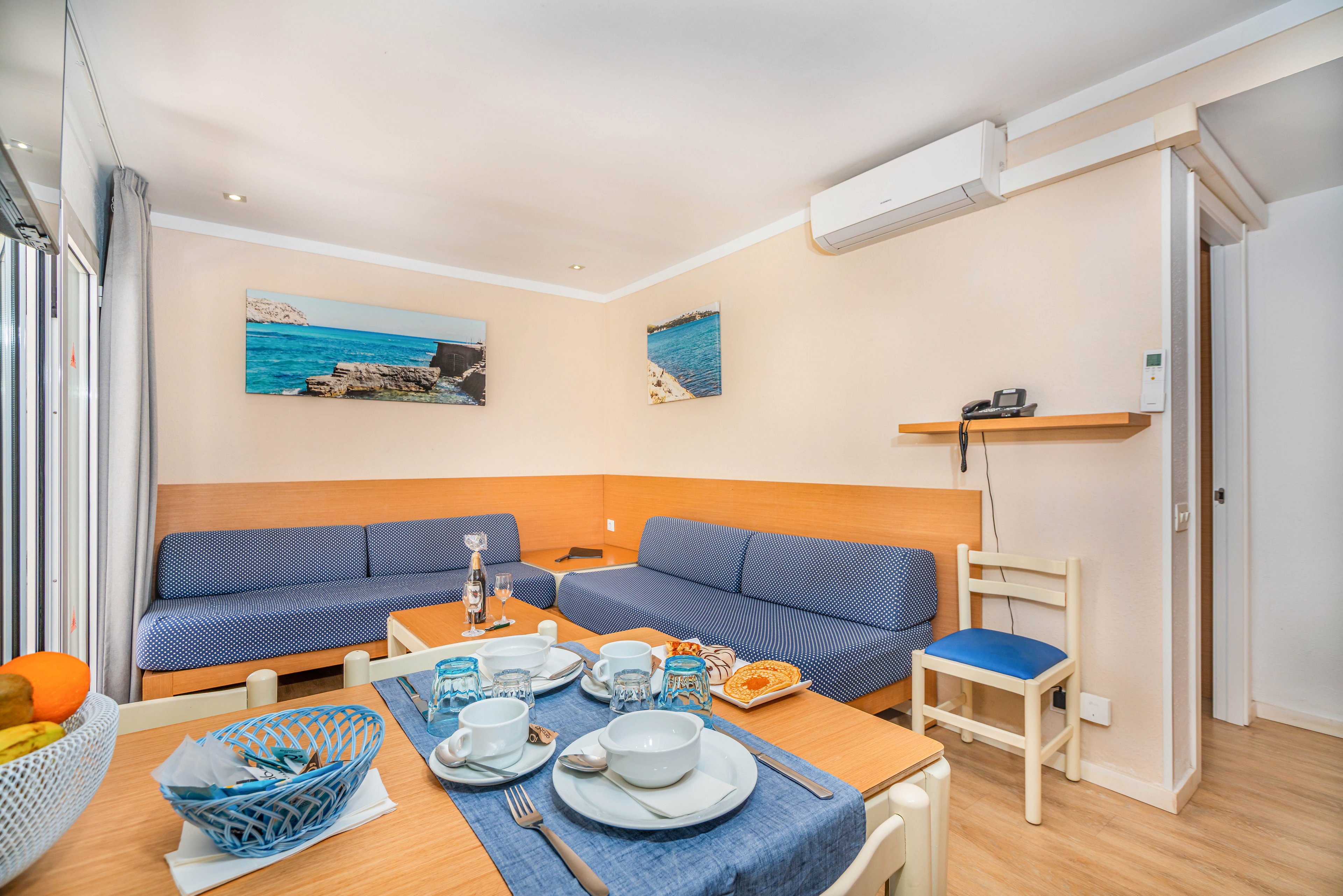 A compact living room features two blue sofas along light wooden walls, creating a cozy seating area. In the foreground, a small dining table is set with white dishes, cups, and a blue placemat, suggesting a casual meal. Wall art depicting coastal scenes and a bowl of fruit add color and a seaside theme to the space. The room is brightly lit with natural light and includes a mounted air conditioning unit for comfort.