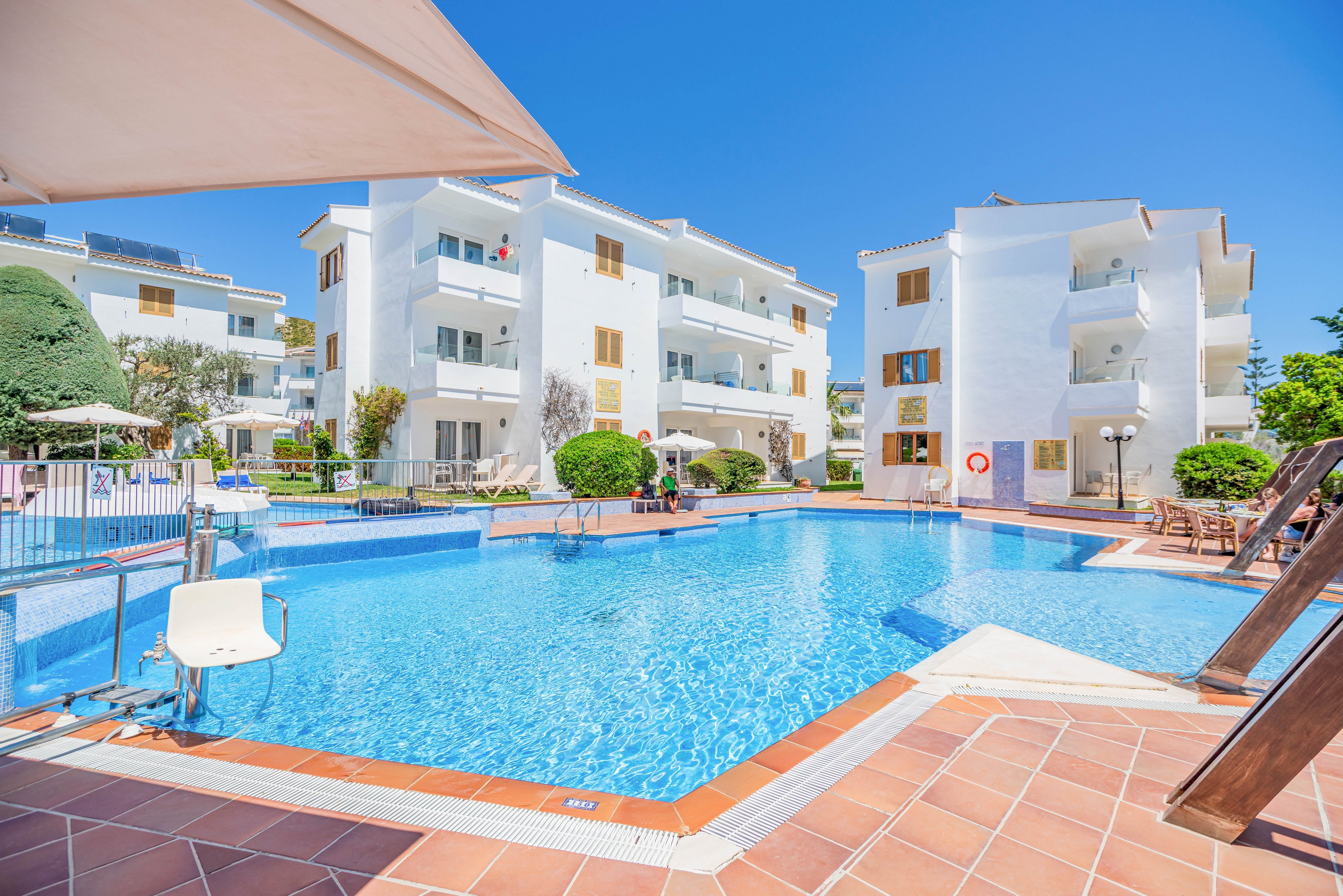 A bright outdoor swimming pool is surrounded by modern white apartment buildings under a clear blue sky. The pool area features tiled decking, metal railings, and a pool lift chair for accessibility. Green shrubs and small trees add contrast to the white facades and blue water. Lounge chairs and umbrellas suggest a relaxing vacation or resort setting.