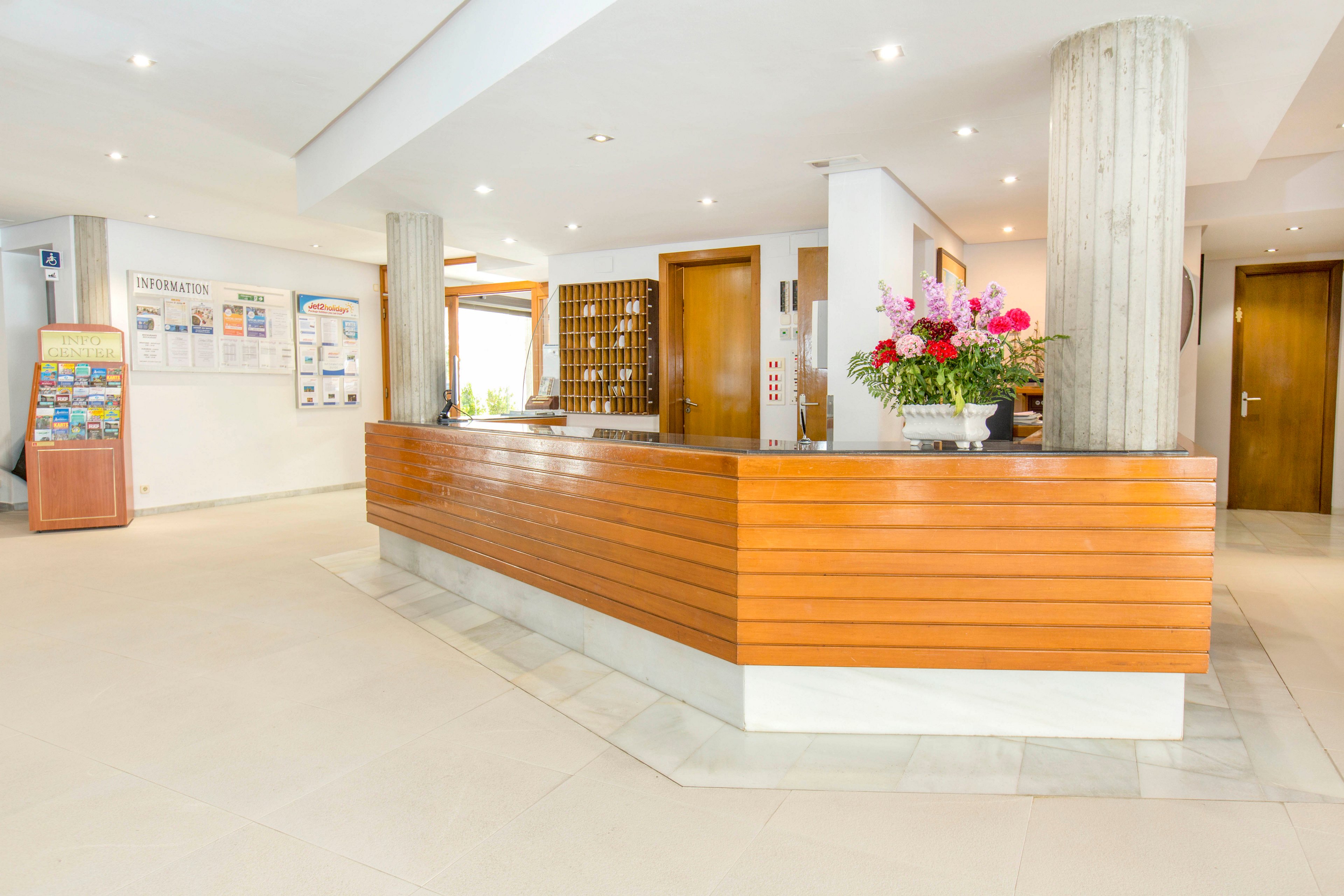 A modern hotel reception area features a long wooden front desk with clean, angular lines. The space is bright and minimal, with light-colored floors, white walls, and recessed ceiling lights. Behind the counter are wooden doors, a key rack, and informational boards on the wall. A large floral arrangement adds color and warmth to the otherwise neutral lobby. The overall atmosphere is tidy, welcoming, and professional.