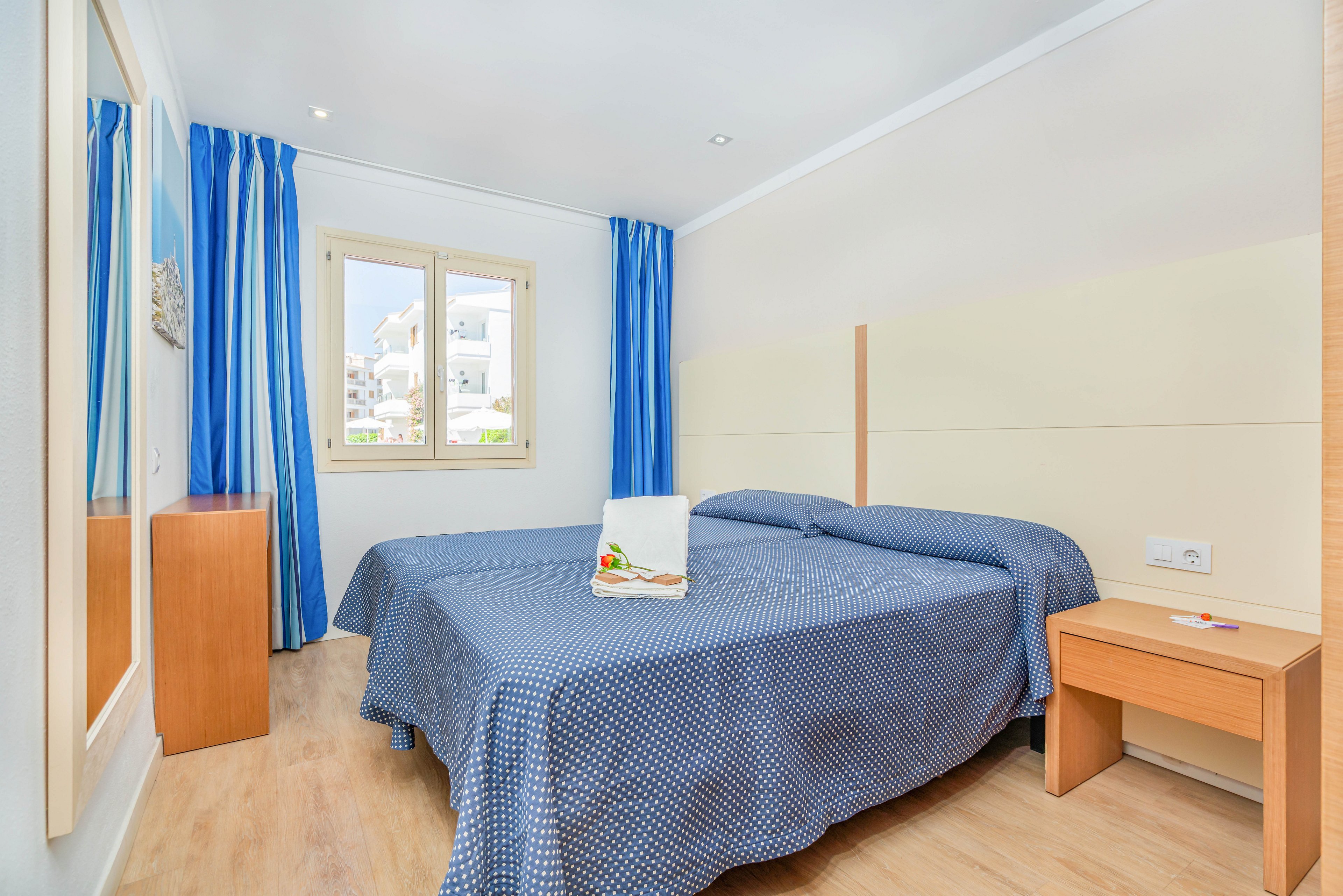 A clean, modern bedroom features two twin beds pushed together, covered with blue patterned bedspreads. The room has light wooden furniture, including a bedside table and a small desk or dresser. Blue curtains frame a window that lets in natural daylight and reveals neighboring buildings outside. A neatly folded white towel with a small decorative arrangement rests on one of the beds, adding a welcoming touch.