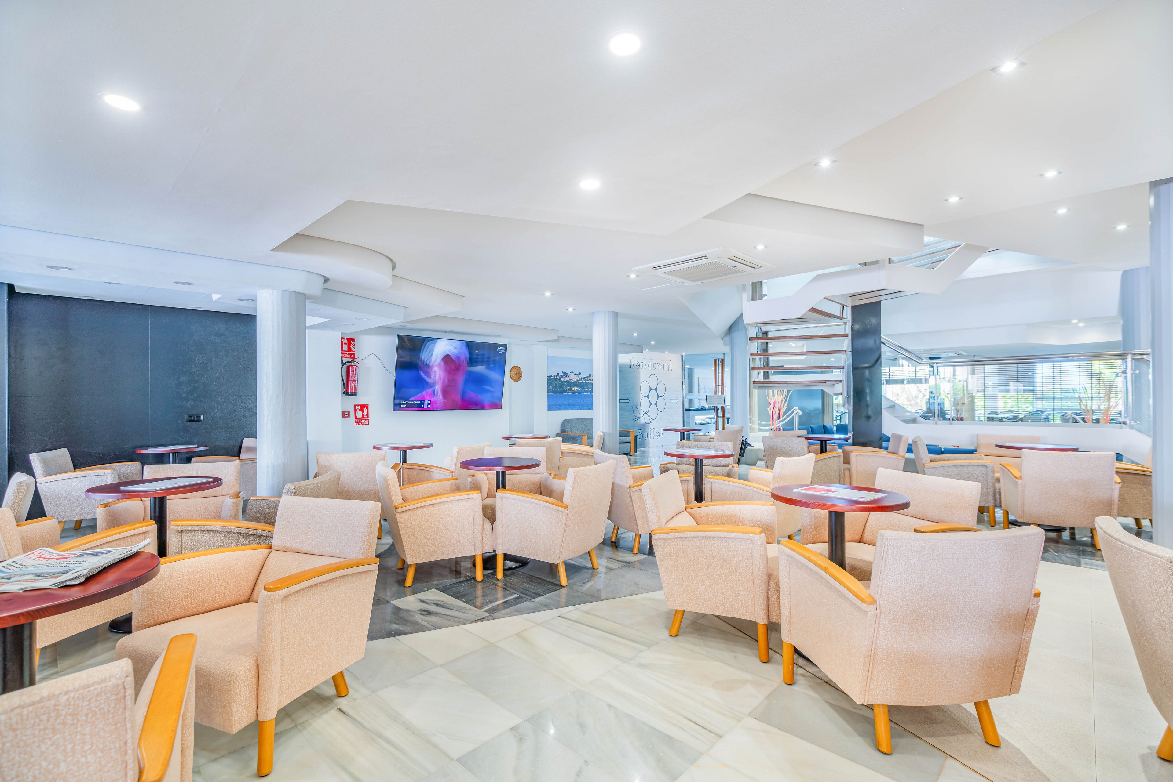 A spacious, modern lounge area features numerous beige armchairs arranged around small round wooden tables. The setting appears to be a hotel or café with a polished floor, white walls, and recessed ceiling lights creating a bright atmosphere. A large flat-screen television is mounted on the wall, and a spiral staircase is visible in the background. The room is empty of people, emphasizing the clean, orderly layout and contemporary design.