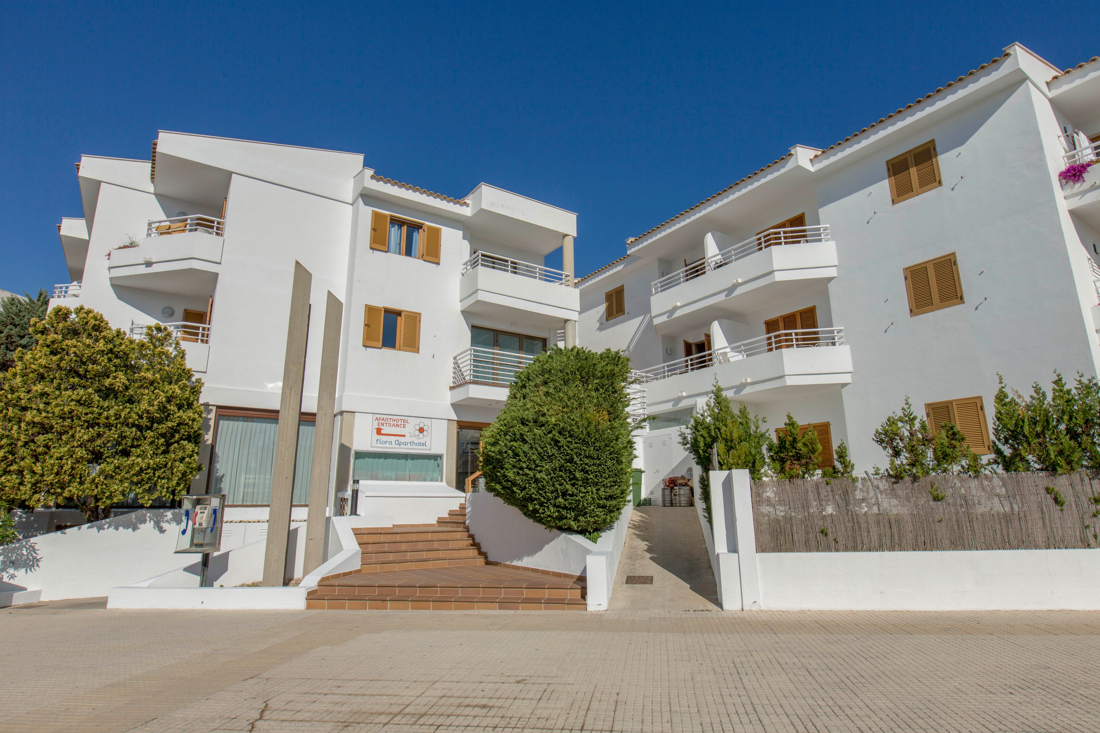 A modern white apartment complex is shown under a clear blue sky. The buildings feature multiple balconies, wooden shutters, and clean geometric lines. A central staircase leads up from a paved courtyard, framed by trimmed bushes and trees. The bright sunlight and absence of people create a calm, quiet residential atmosphere.