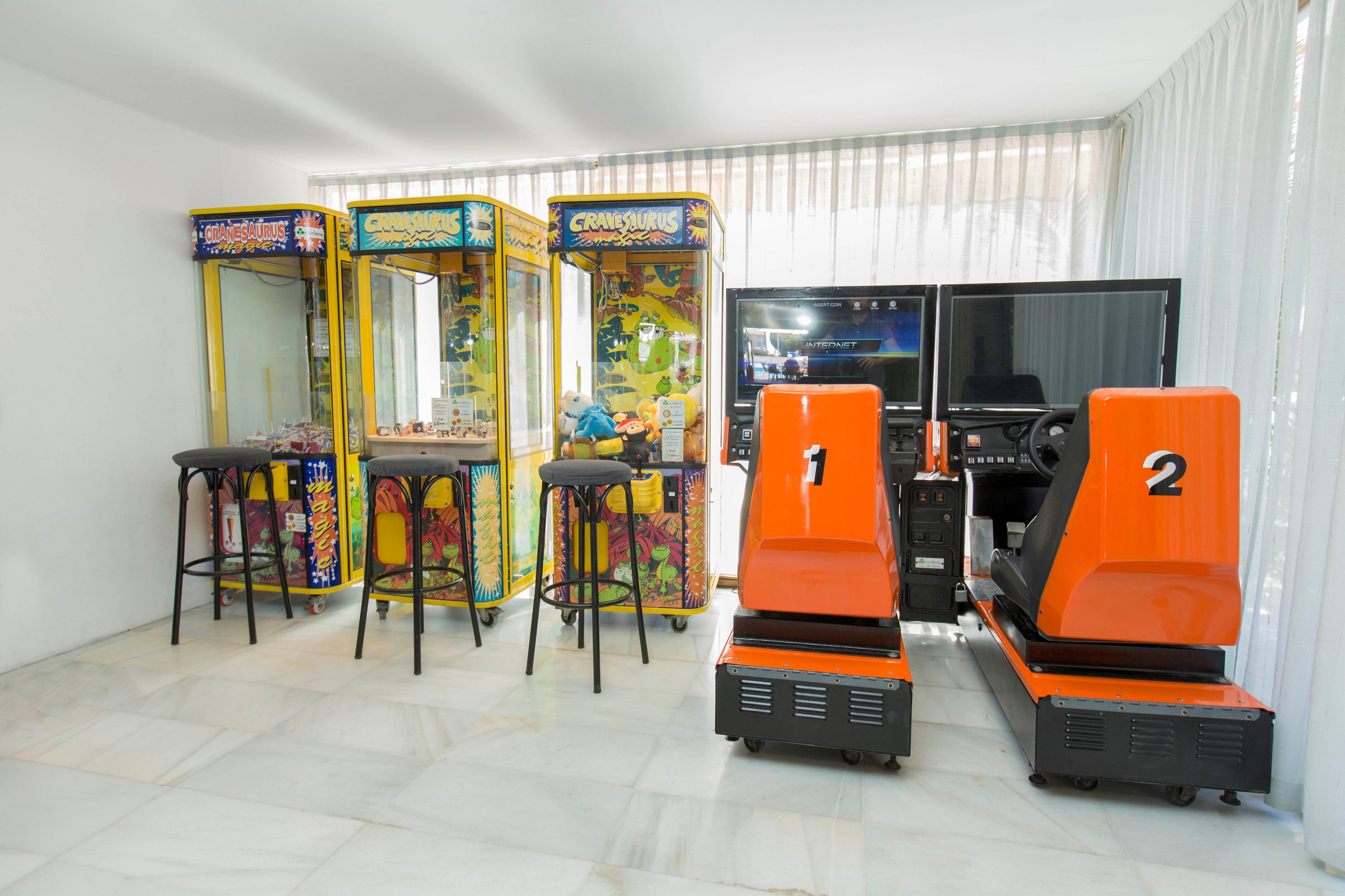 A bright arcade-style game room features several claw machines and two orange racing simulators. The machines are lined up against a white wall with sheer curtains allowing natural light to filter in. High stools are placed in front of the claw machines, suggesting a casual play area. The overall scene feels clean, minimal, and modern with a focus on entertainment equipment.