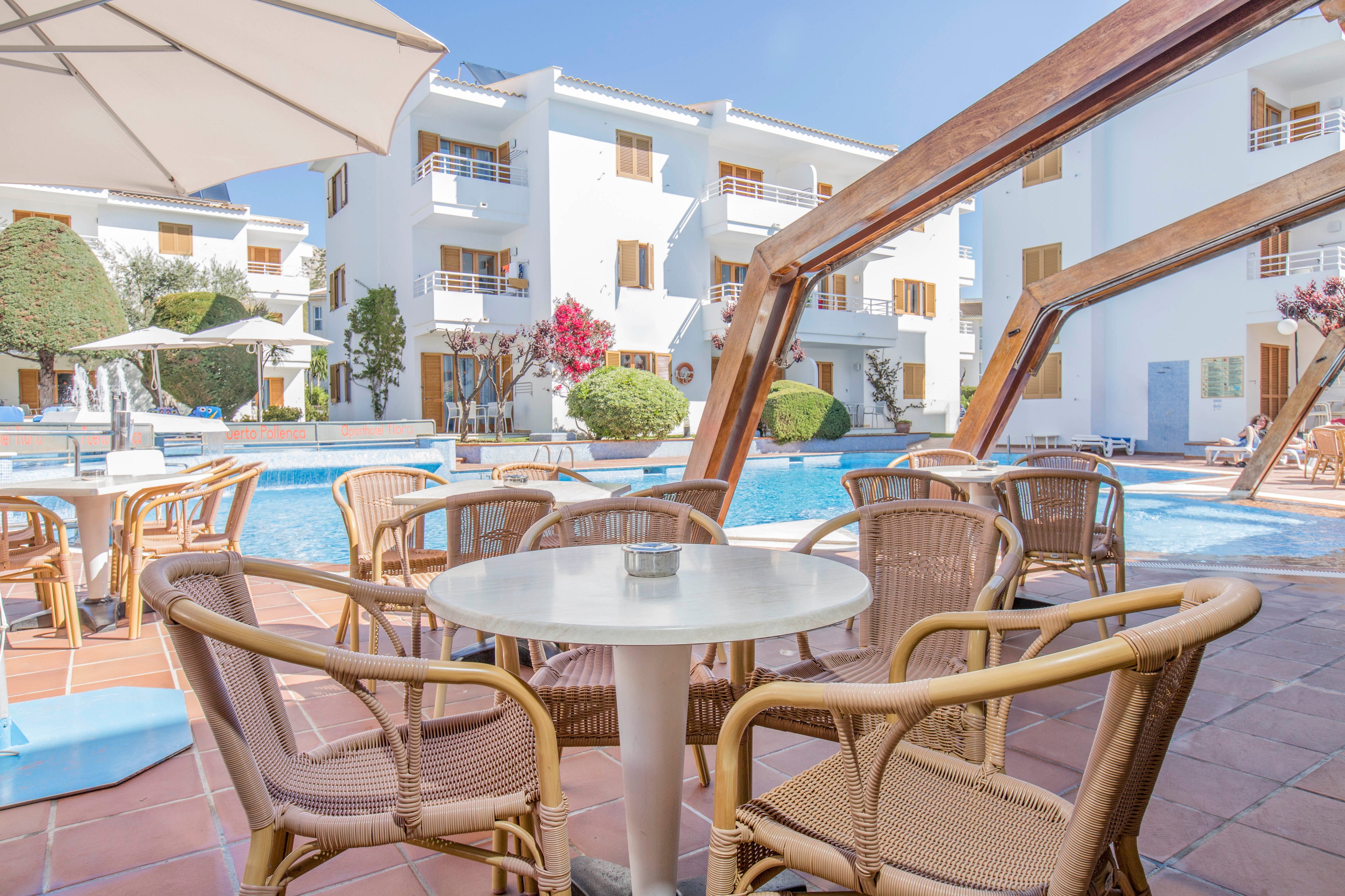 An outdoor seating area with wicker chairs and a round white table overlooks a bright blue swimming pool. White multi-story buildings with balconies surround the courtyard, creating a resort-like atmosphere. Trimmed trees, flowering shrubs, and poolside umbrellas add greenery and shade to the sunny scene. The clear sky and calm water suggest a warm, relaxing vacation setting.