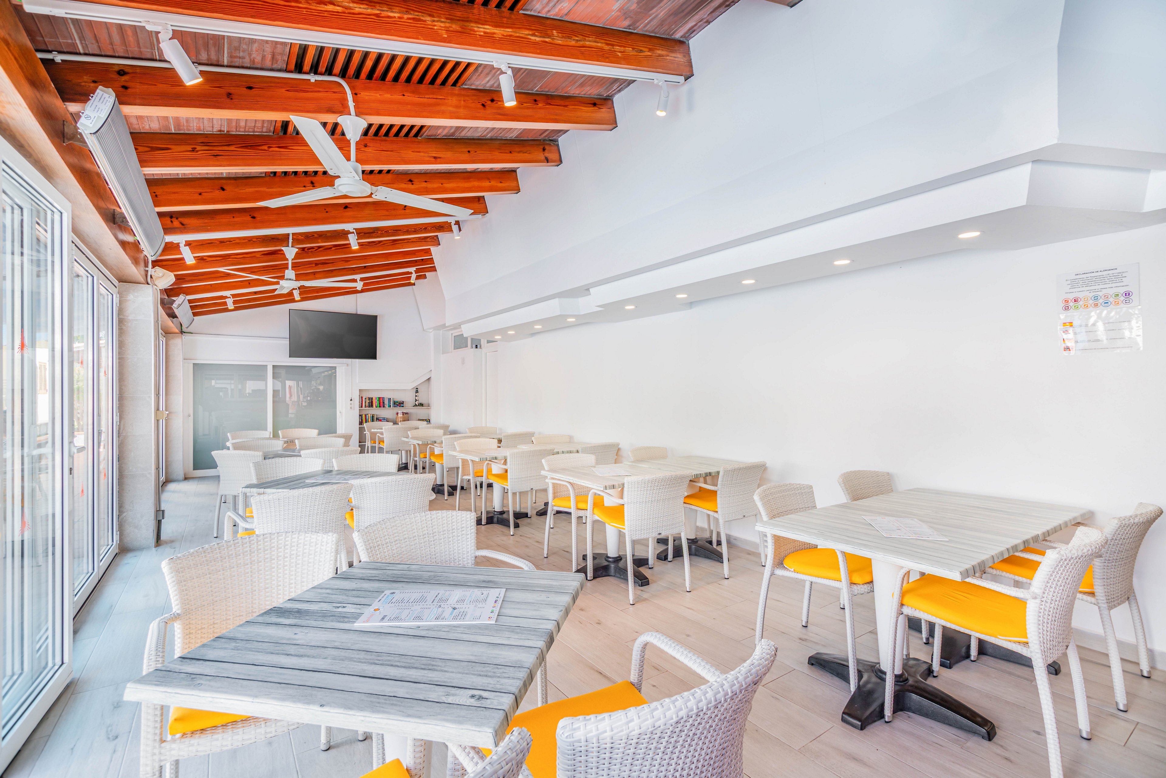 A bright modern cafe interior features white wicker chairs with yellow cushions and light wooden tables. The space has a high ceiling with exposed wooden beams and ceiling fans. Large glass doors on one side allow natural light to flood the room, creating an airy atmosphere. A wall-mounted television and recessed ceiling lights add contemporary touches to the clean, minimalist design.