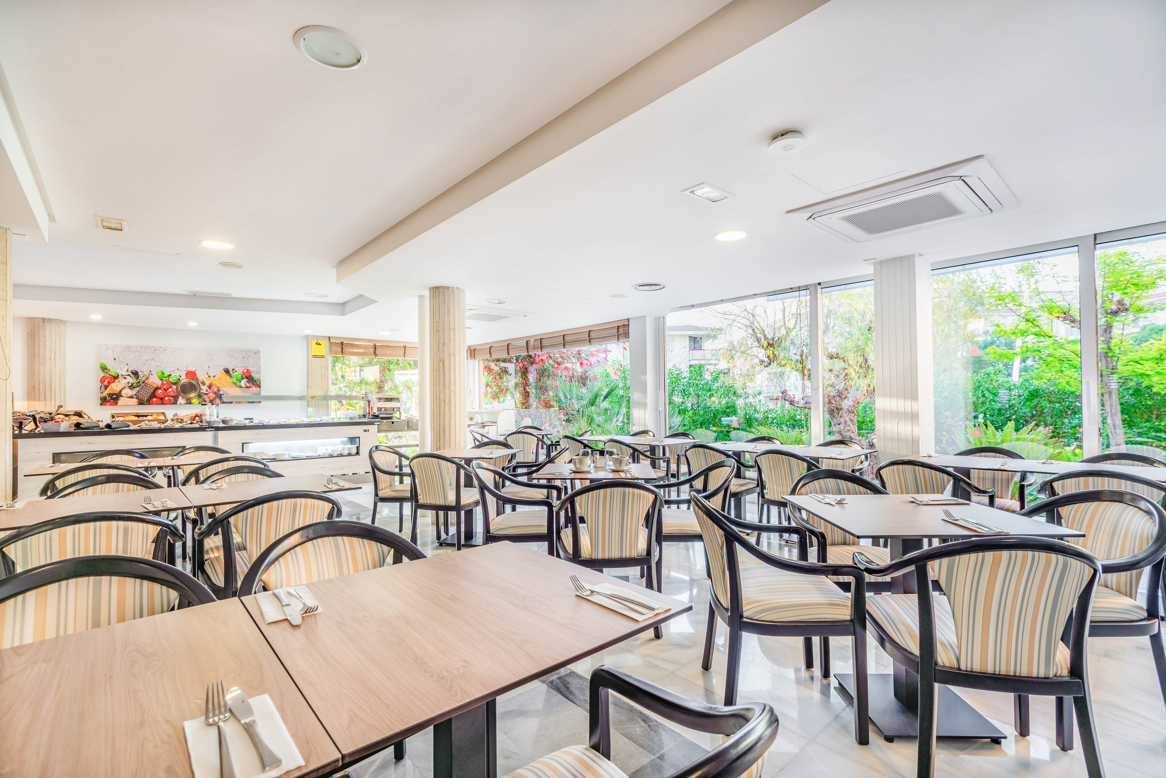 A spacious dining room features neatly arranged wooden tables and striped chairs. Place settings with cutlery and napkins are prepared on each table, suggesting readiness for guests. Large windows along one side let in abundant natural light and reveal a lush green garden outside. In the background, a buffet counter with food displays adds to the atmosphere of a hotel or resort restaurant.