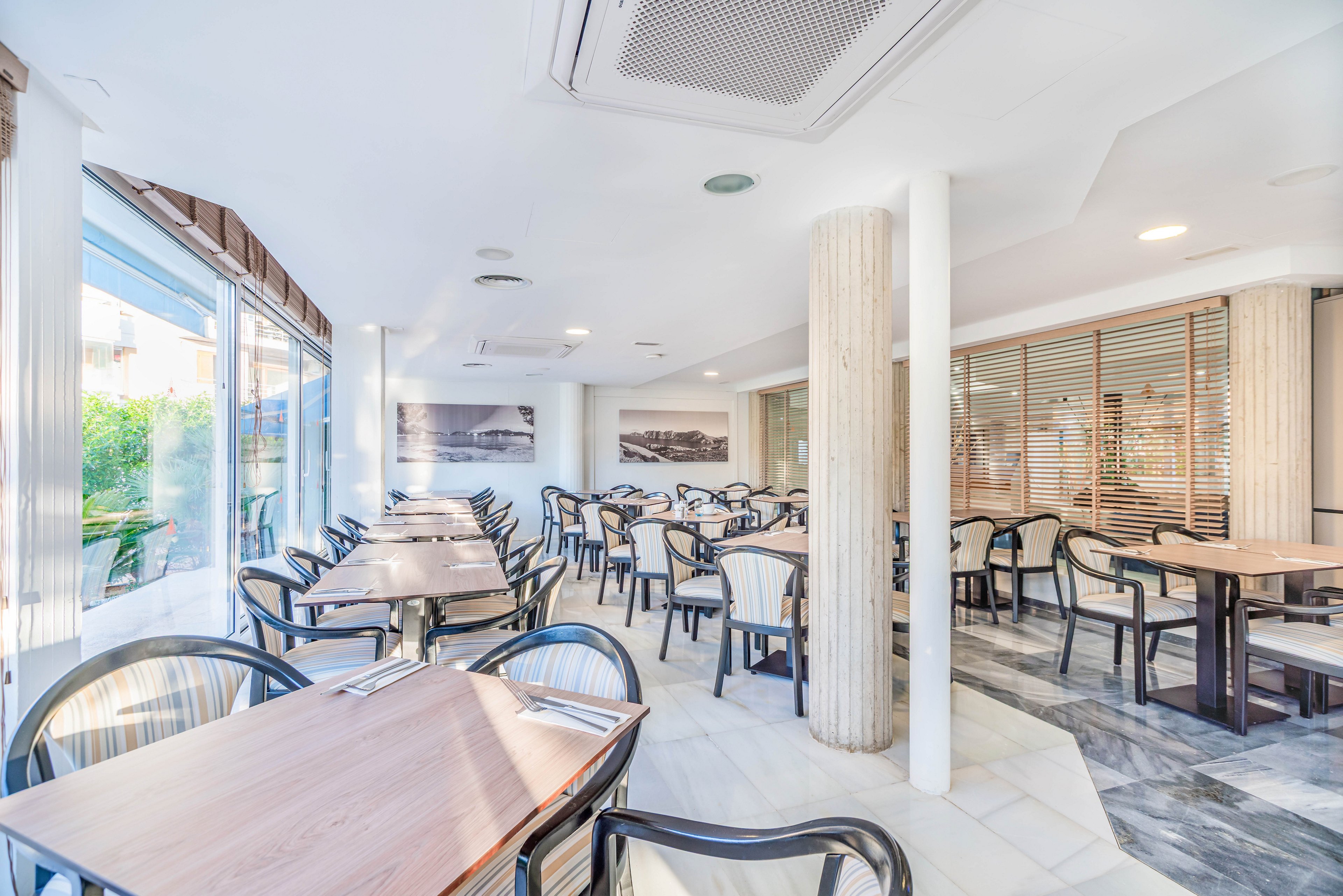 A bright, modern restaurant dining room is shown empty and ready for guests. Rows of wooden tables with black plastic chairs are neatly arranged across the white tiled floor. Large windows on one side let in abundant natural light and offer a view of greenery outside. Neutral tones, clean lines, and wall art create a calm, contemporary atmosphere suitable for casual dining.