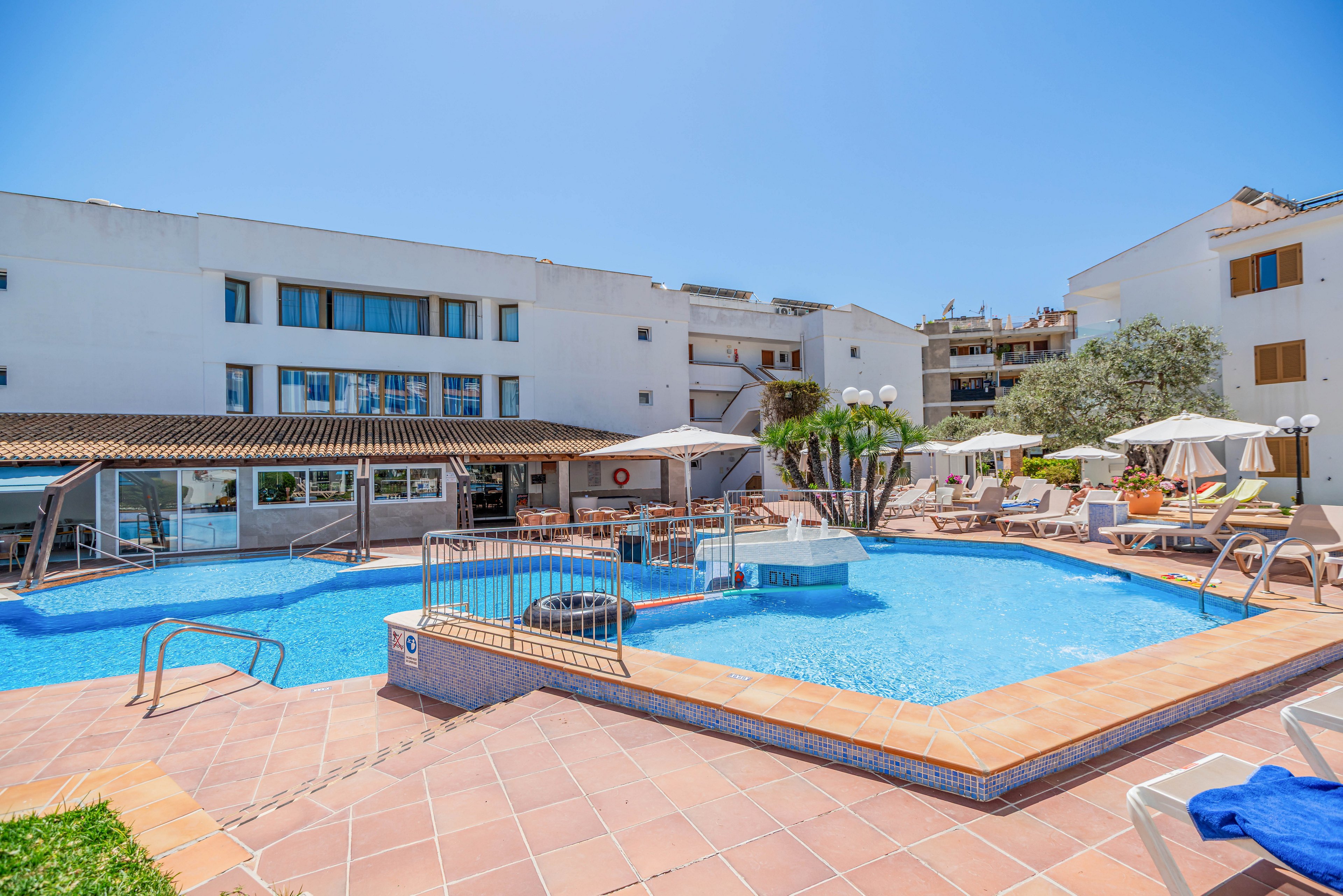 An outdoor hotel swimming pool is surrounded by sun loungers and umbrellas under a clear blue sky. The modern white building forms the backdrop, with balconies overlooking the pool area. Metal railings, tiled pool edges, and a small bridge add structure to the scene. Bright sunlight and clear water create a relaxed vacation atmosphere.