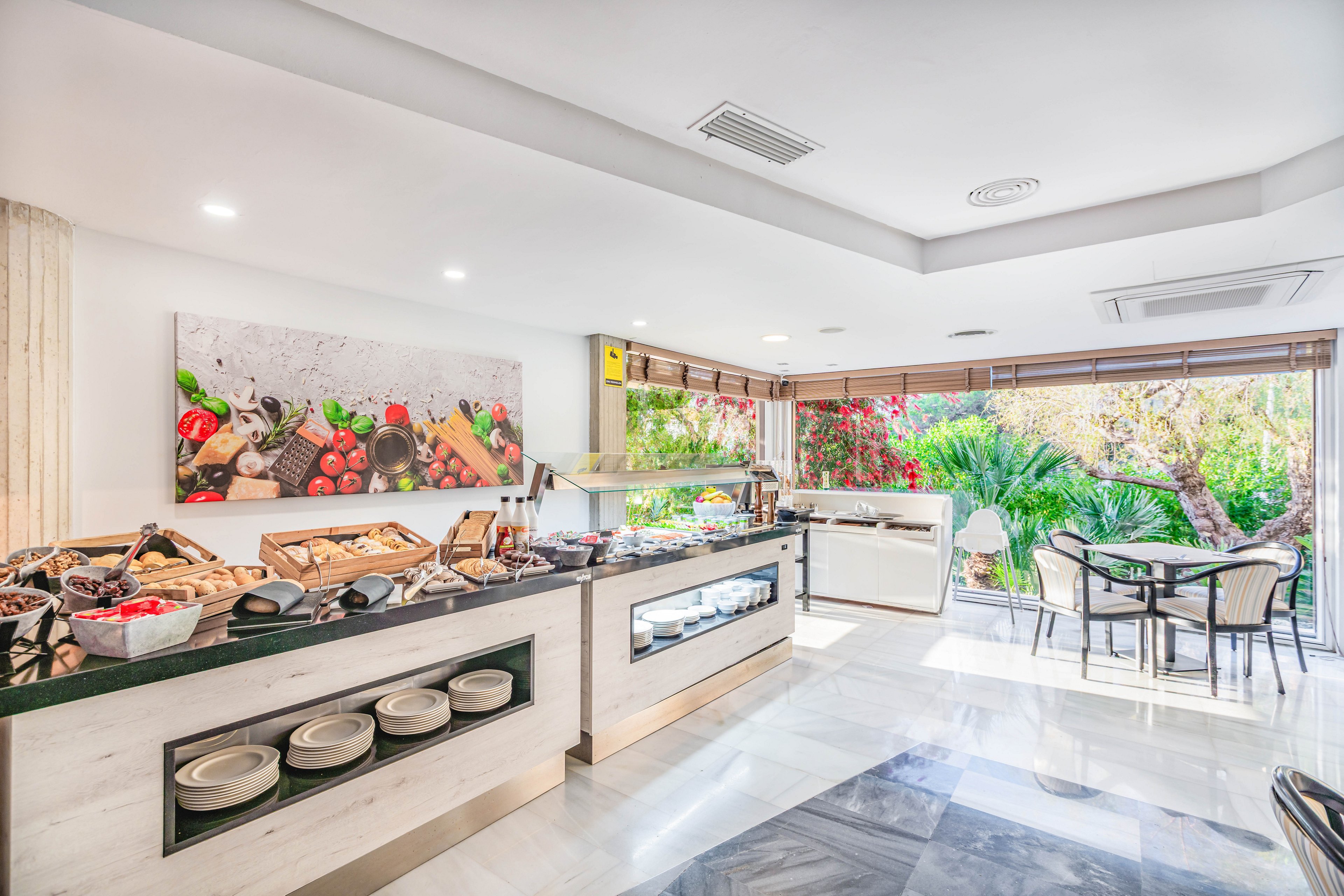 A modern self-service breakfast buffet is arranged on sleek counters in a bright hotel dining area. Various breads, cereals, and cold items are displayed under glass with plates and bowls neatly stored below. Large windows reveal a lush garden with palm trees and flowering plants, bringing natural light into the room. A few tables and chairs are set near the windows, creating a relaxed and inviting atmosphere for guests.