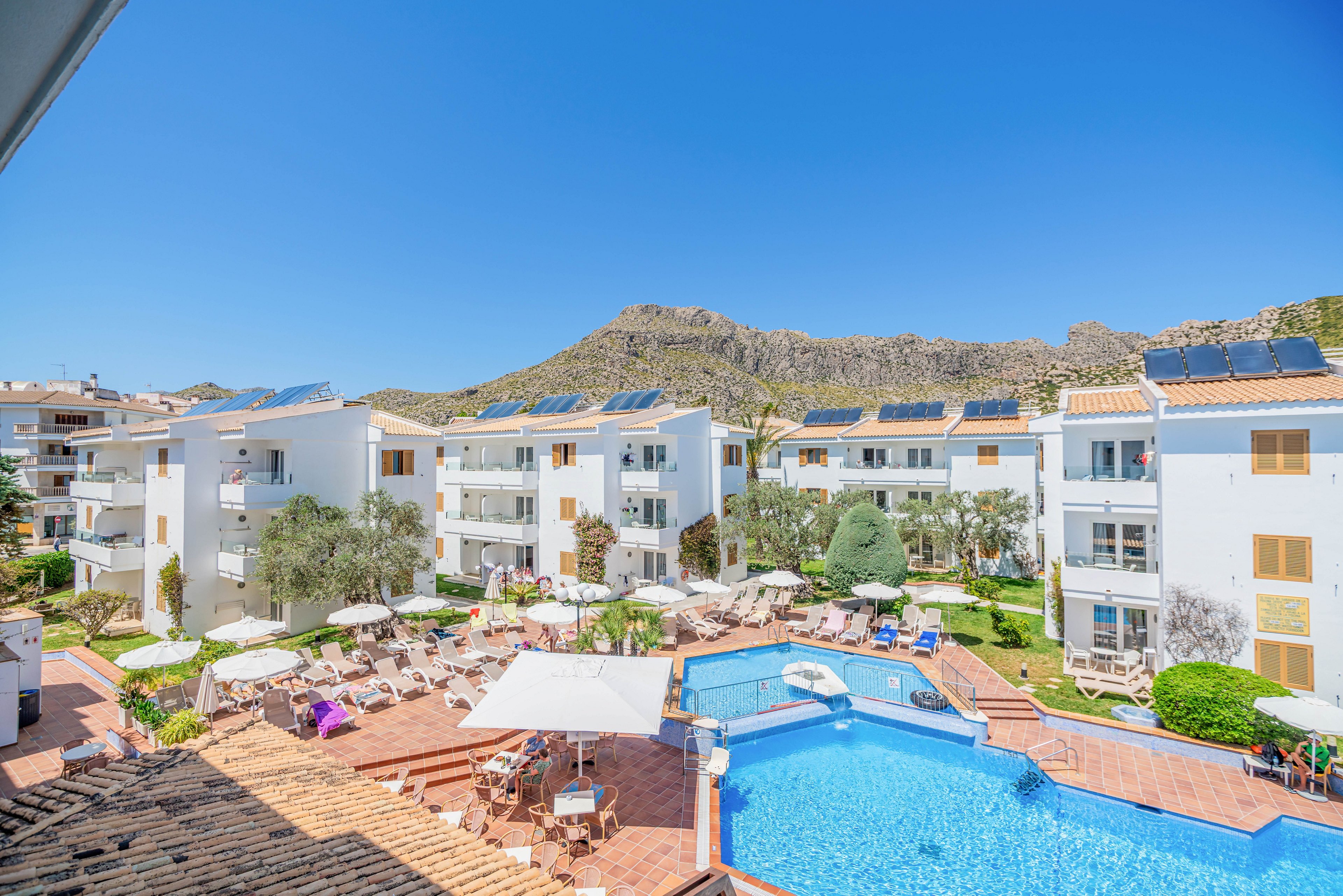 A bright resort complex features several white apartment buildings surrounding a large blue swimming pool. Guests relax on sun loungers and under umbrellas on the poolside terrace. Solar panels are visible on many of the rooftops, and the scene is framed by rugged mountains in the background. The clear blue sky and lush greenery create a warm, inviting holiday atmosphere.