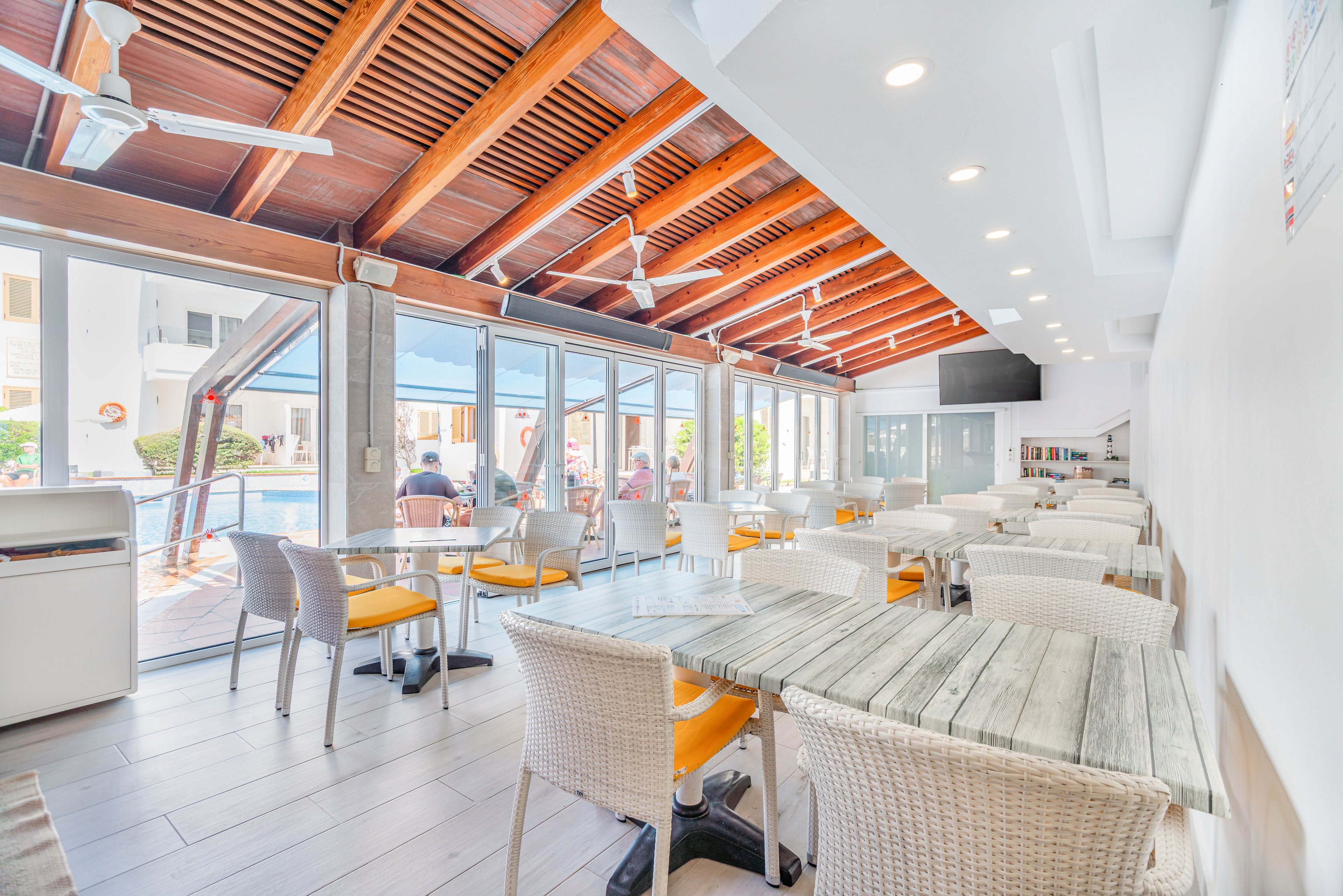 A bright, modern cafe interior features light wooden tables and wicker chairs with yellow cushions. Large floor-to-ceiling glass doors open to an outdoor pool area where people are seated in the background. The space has a high wooden ceiling with exposed beams, ceiling fans, and recessed lighting. Overall, the scene feels airy, relaxed, and inviting for casual dining or refreshments.