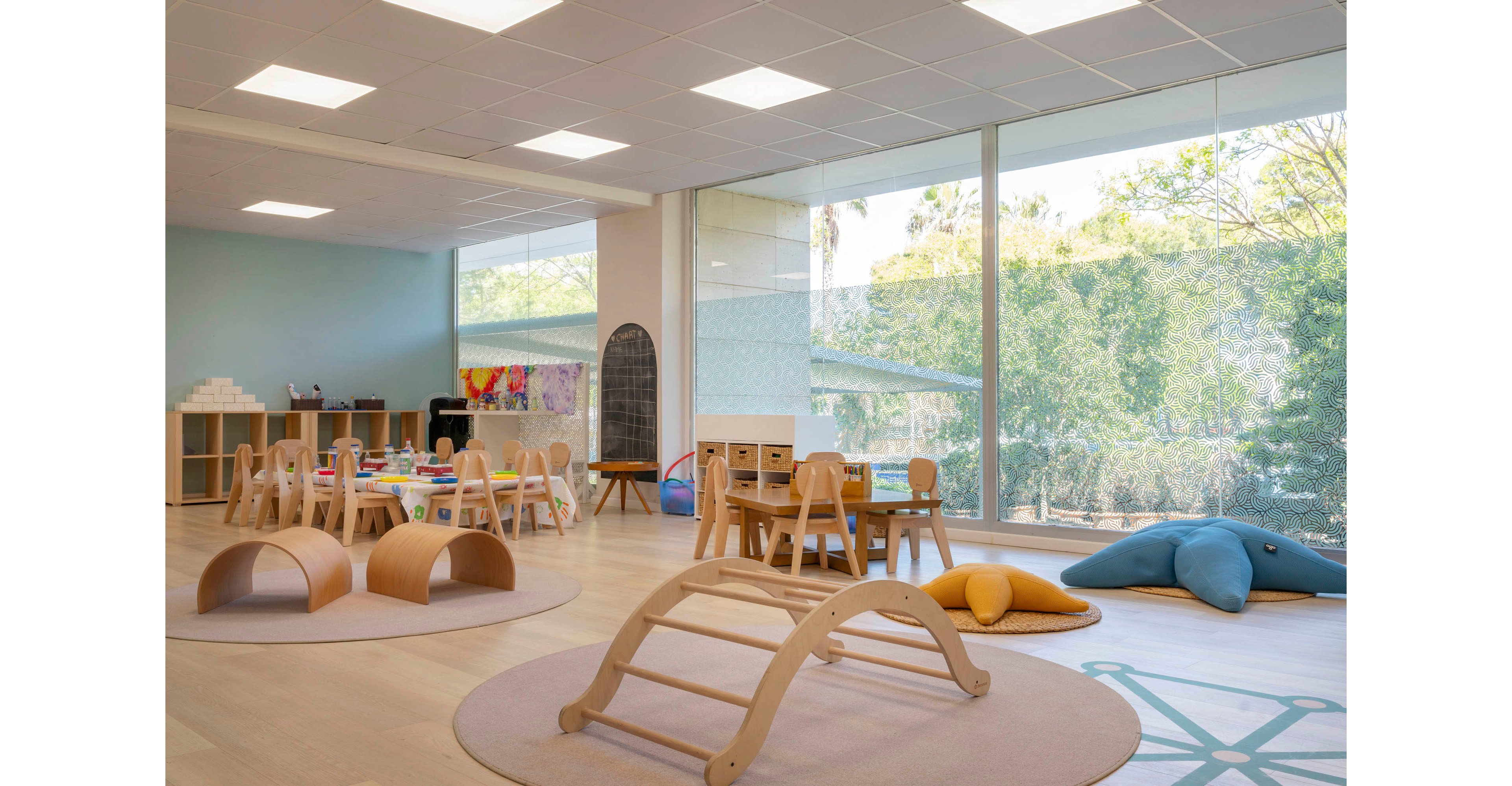 Spacious preschool classroom featuring wooden play structures and child-sized furniture. The room includes low tables, small chairs, and curved climbing frames arranged on round rugs. Soft cushions and floor mats create cozy play and reading zones near large floor-to-ceiling windows. Natural light fills the space, highlighting the minimalist decor and view of trees outside.